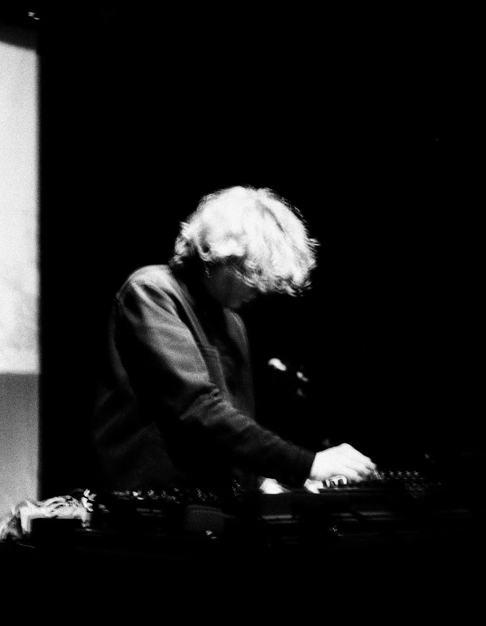 Black and white photo of a person with curly hair playing a keyboard or synthesizer in a dark setting.