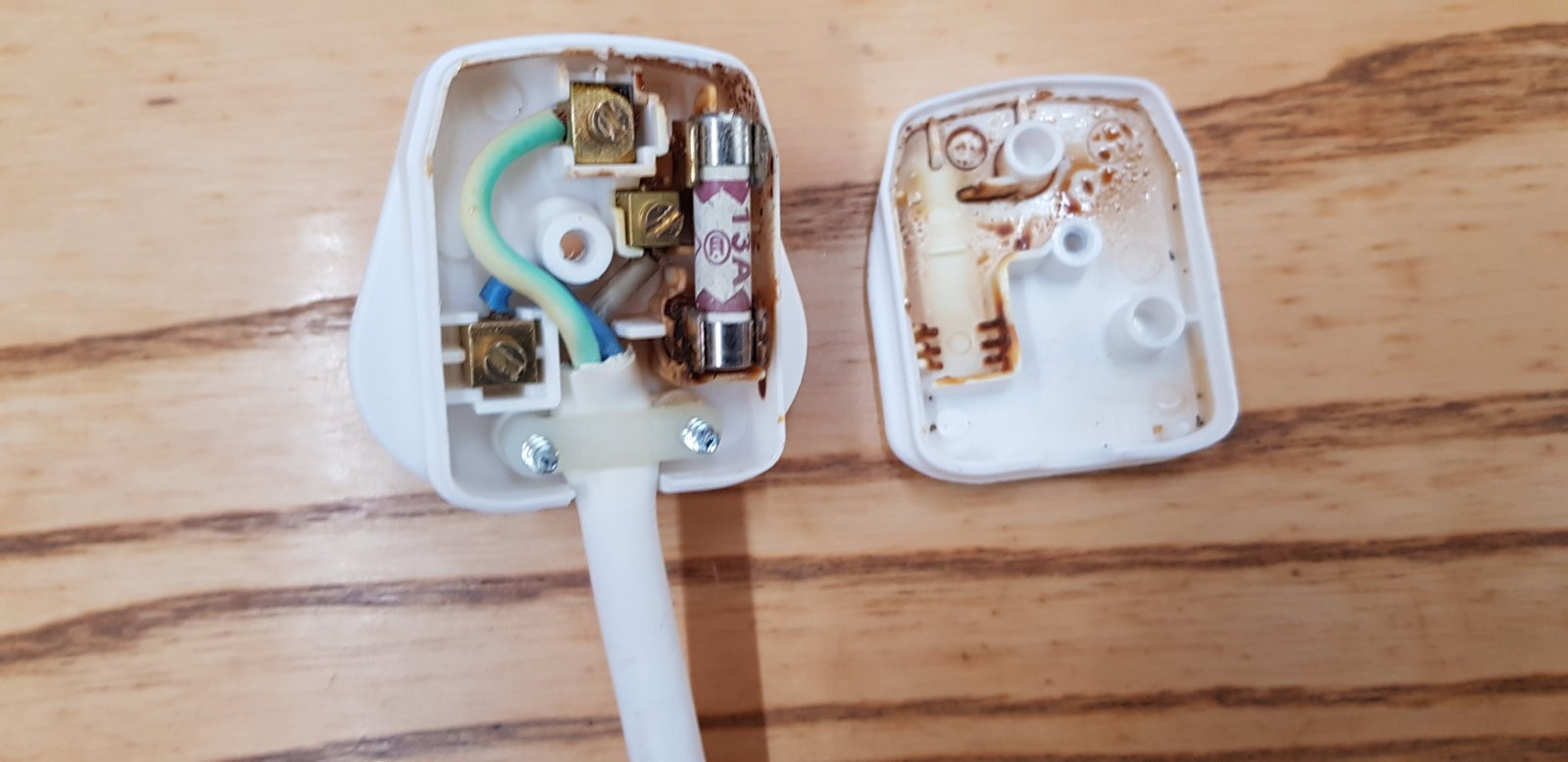 Disassembled electrical power plug with metal prongs, wiring, and the plastic casing on a wooden surface.