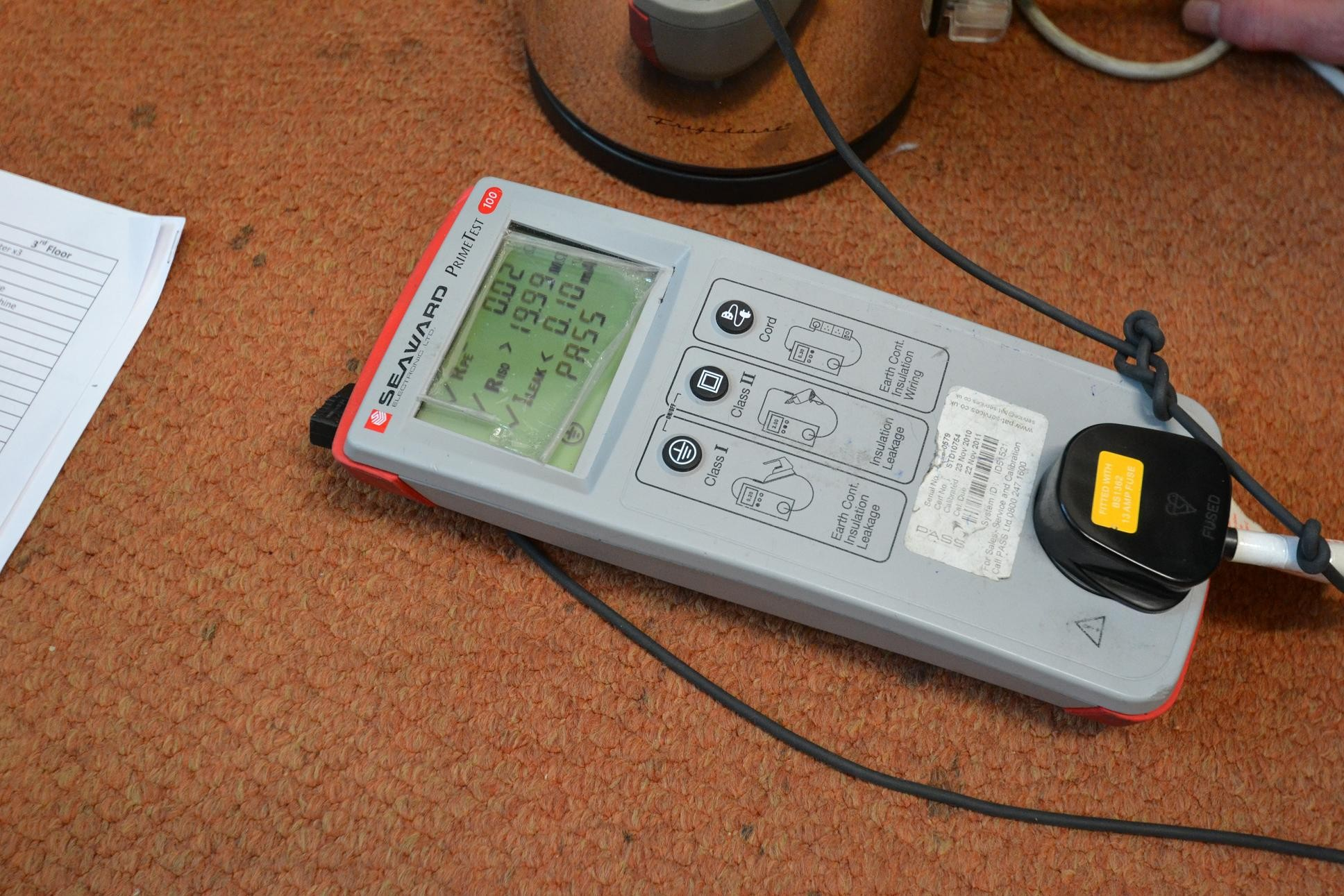 A digital electrical testing device on a carpeted floor with a power cord plugged into it, displaying measurements on its screen.
