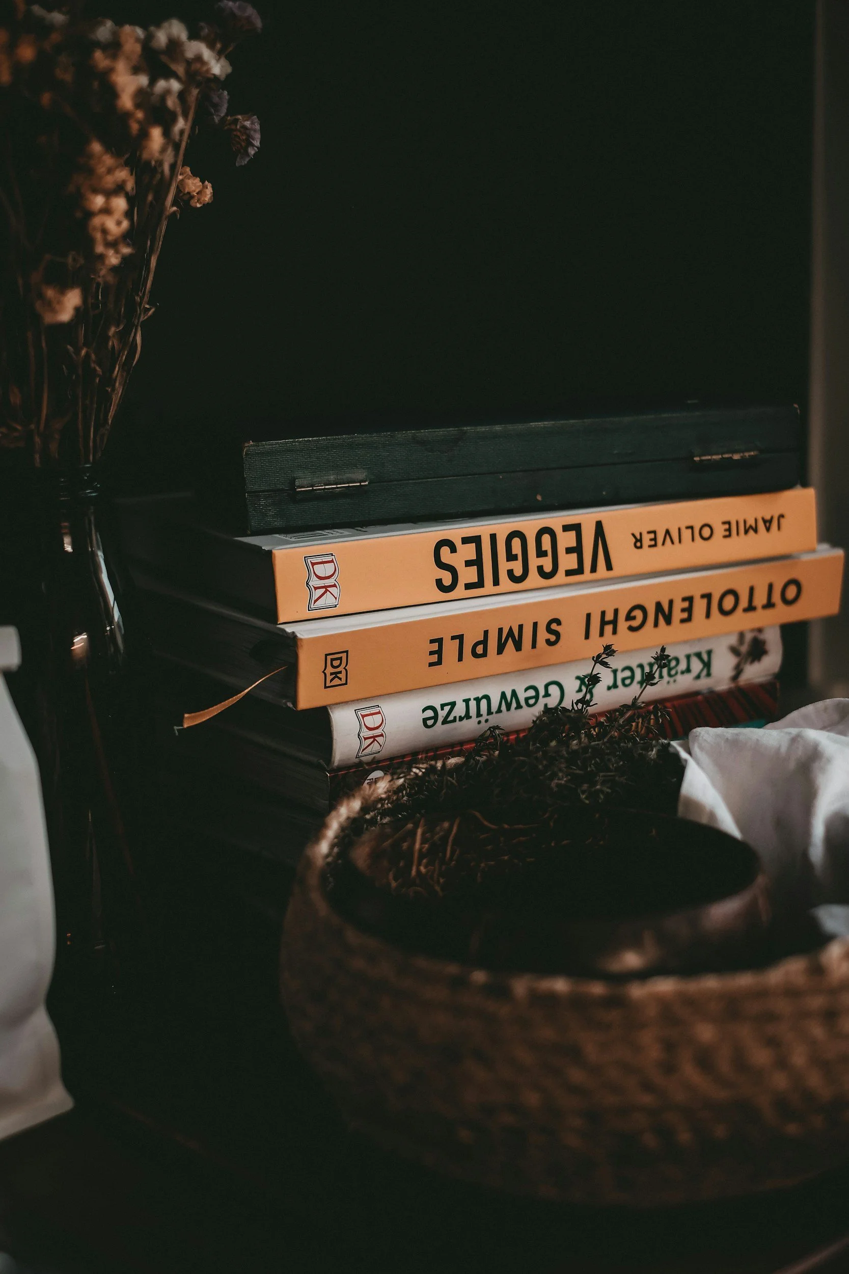 Une pile de livres avec des titres en allemand, entourée de plantes séchées et placée dans un panier en osier, dans une ambiance chaleureuse et sombre.