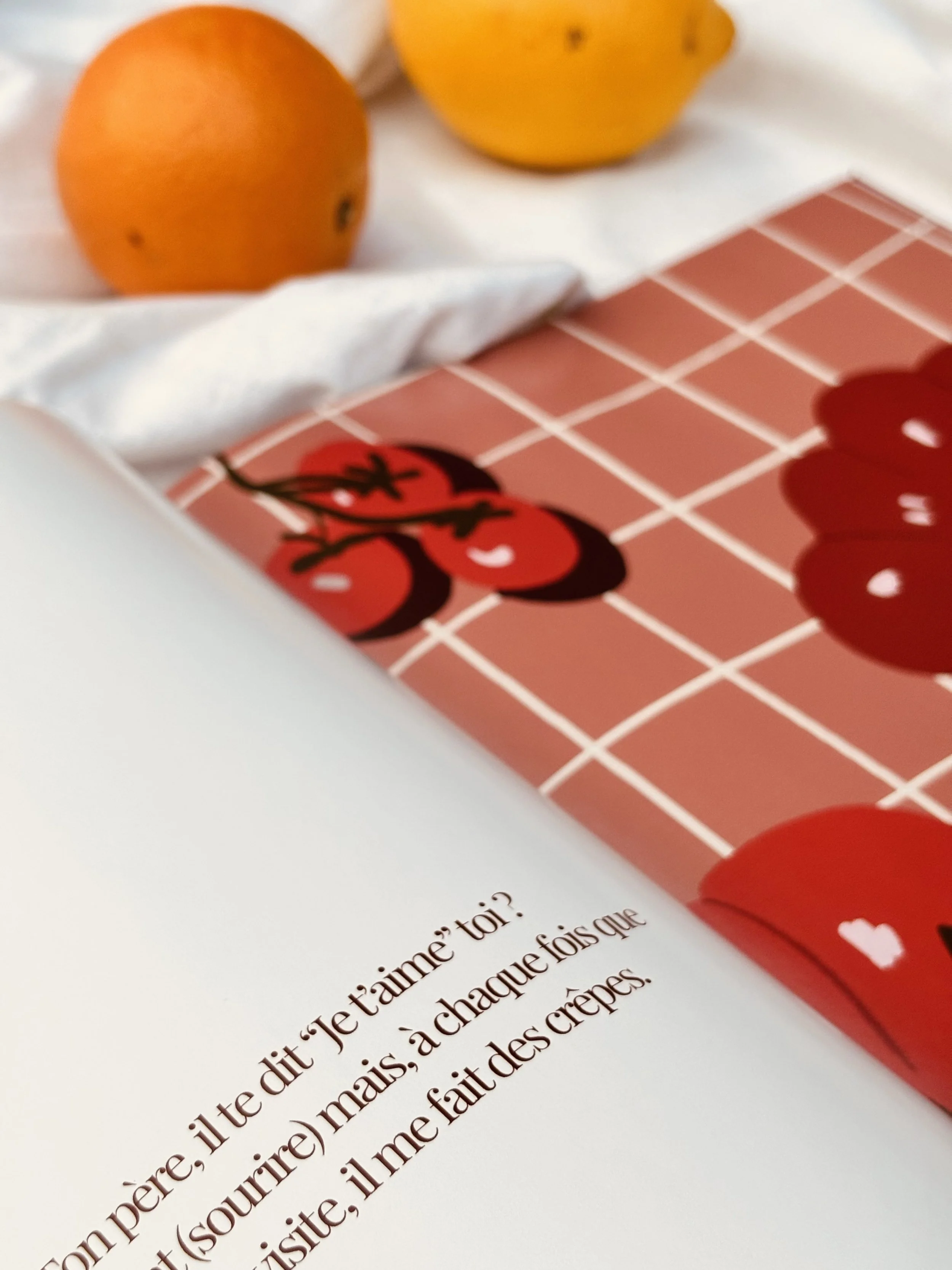 Un livre ouvert avec un texte en français, une illustration de cerises rouges et un fond en carreaux rouges, avec deux oranges en arrière-plan sur un tissu blanc.