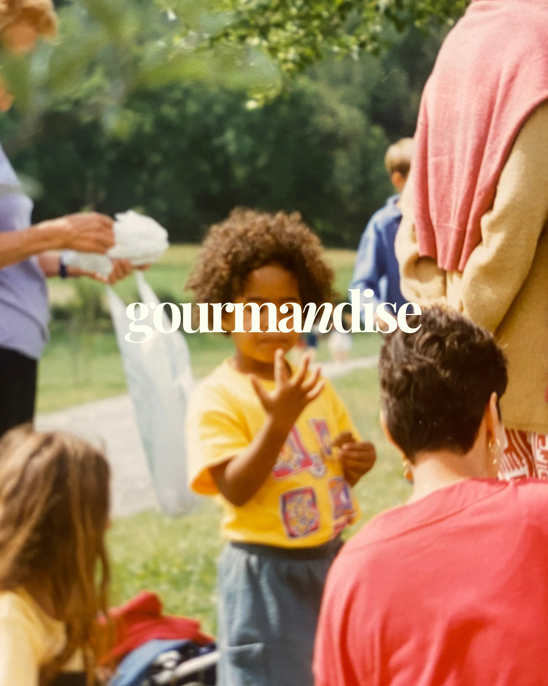 Photo Chez berni : Groupe d'enfants et d'adultes dans un parc lors d'un pique-nique ou d'une fête en plein air, avec un enfant au centre qui mange une pomme.