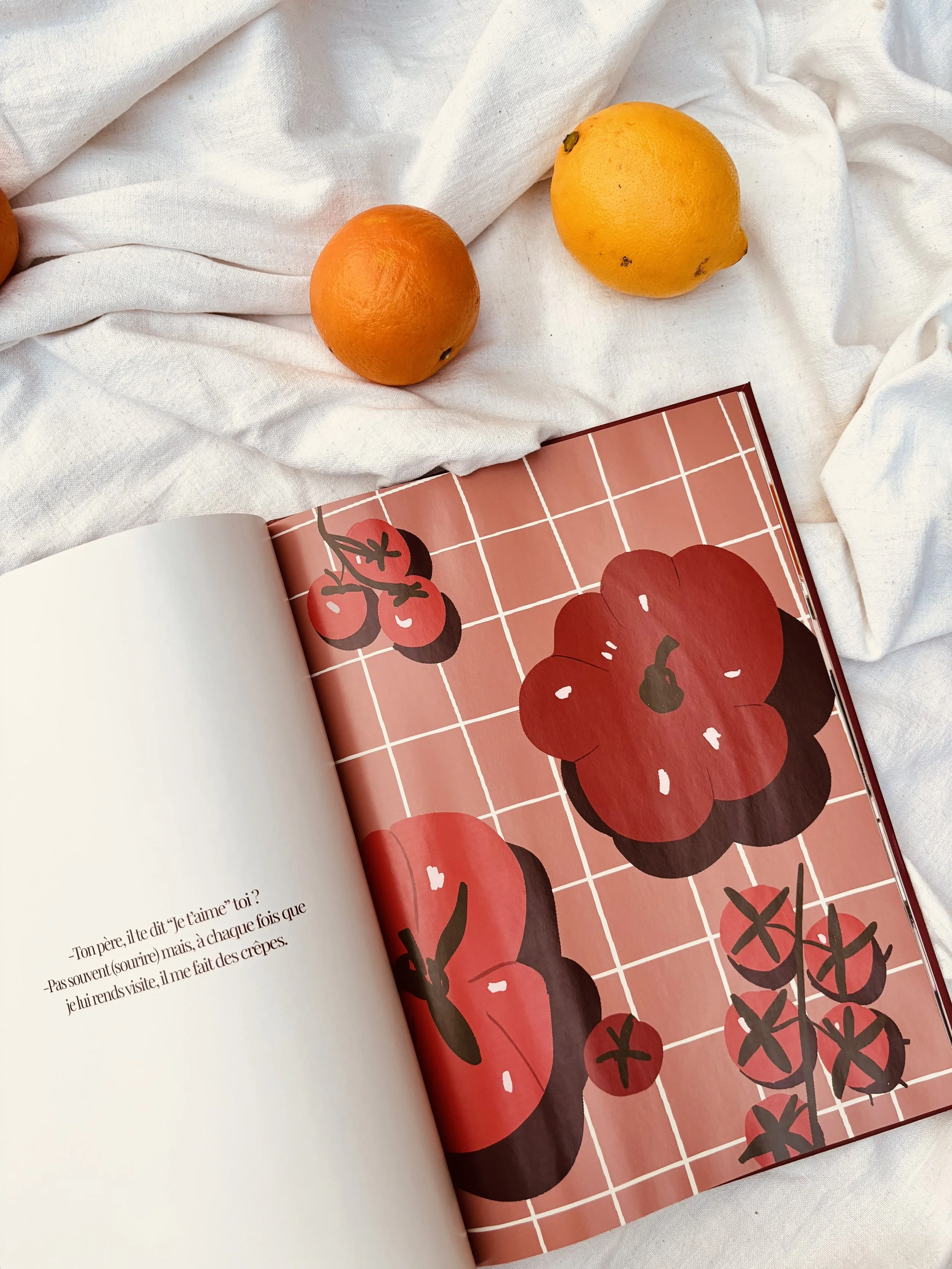 Un livre ouvert avec des illustrations de tomates rouges sur une page et du texte, entouré de deux oranges et un citron posés sur un tissu blanc.