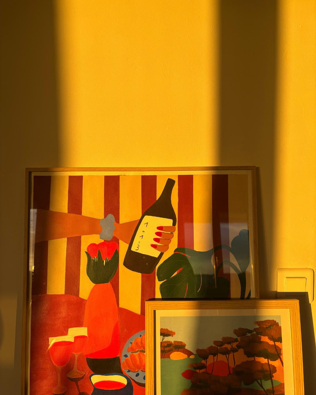 Deux tableaux accrochés sur un mur jaune, l'un représentant une bouteille de vin, une main à ongles rouges, un vase rouge avec une fleur, et des objets culinaires; l'autre représentant une scène de nature avec des arbres et un coucher de soleil.