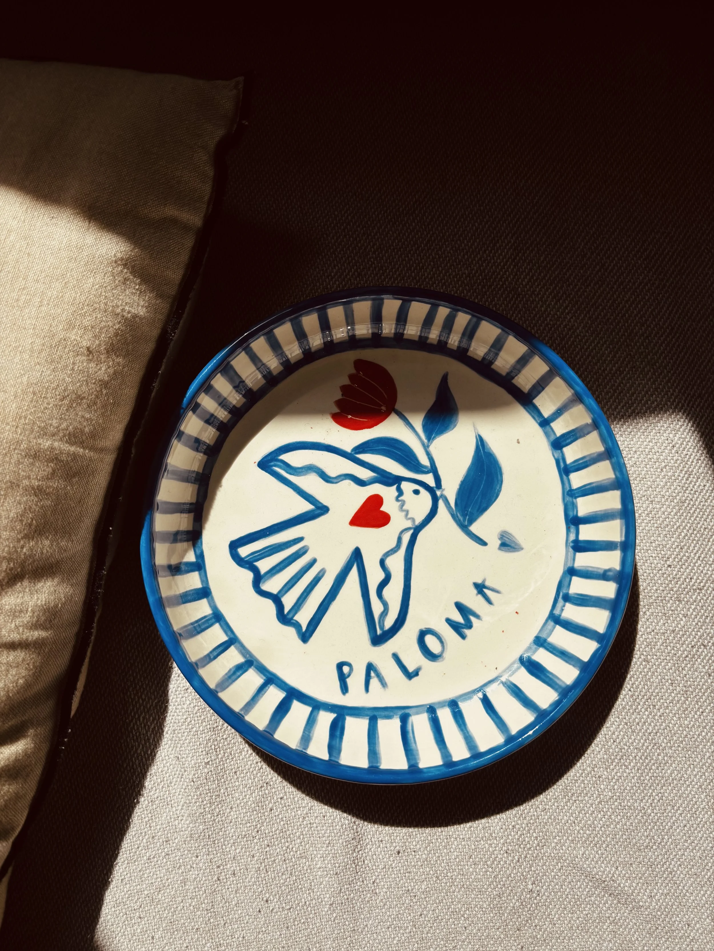 Assiette en céramique décorée de façon colorée avec un dessin d'une colombe, un cœur rouge, une fleur, les mots 'PALOMA', et un fond blanc avec un bord bleu et blanc.