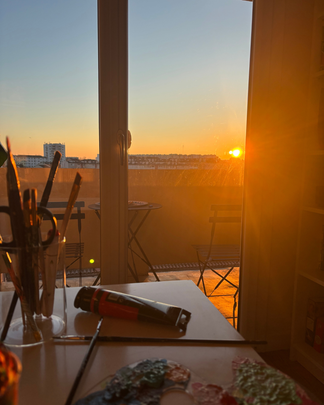 Vue d'une pièce avec un balcon, des tablettes d'artistes et un tube de peinture sur la table, au crépuscule avec un soleil orange à l'horizon, lumière chaude pénétrant à travers la grande fenêtre.