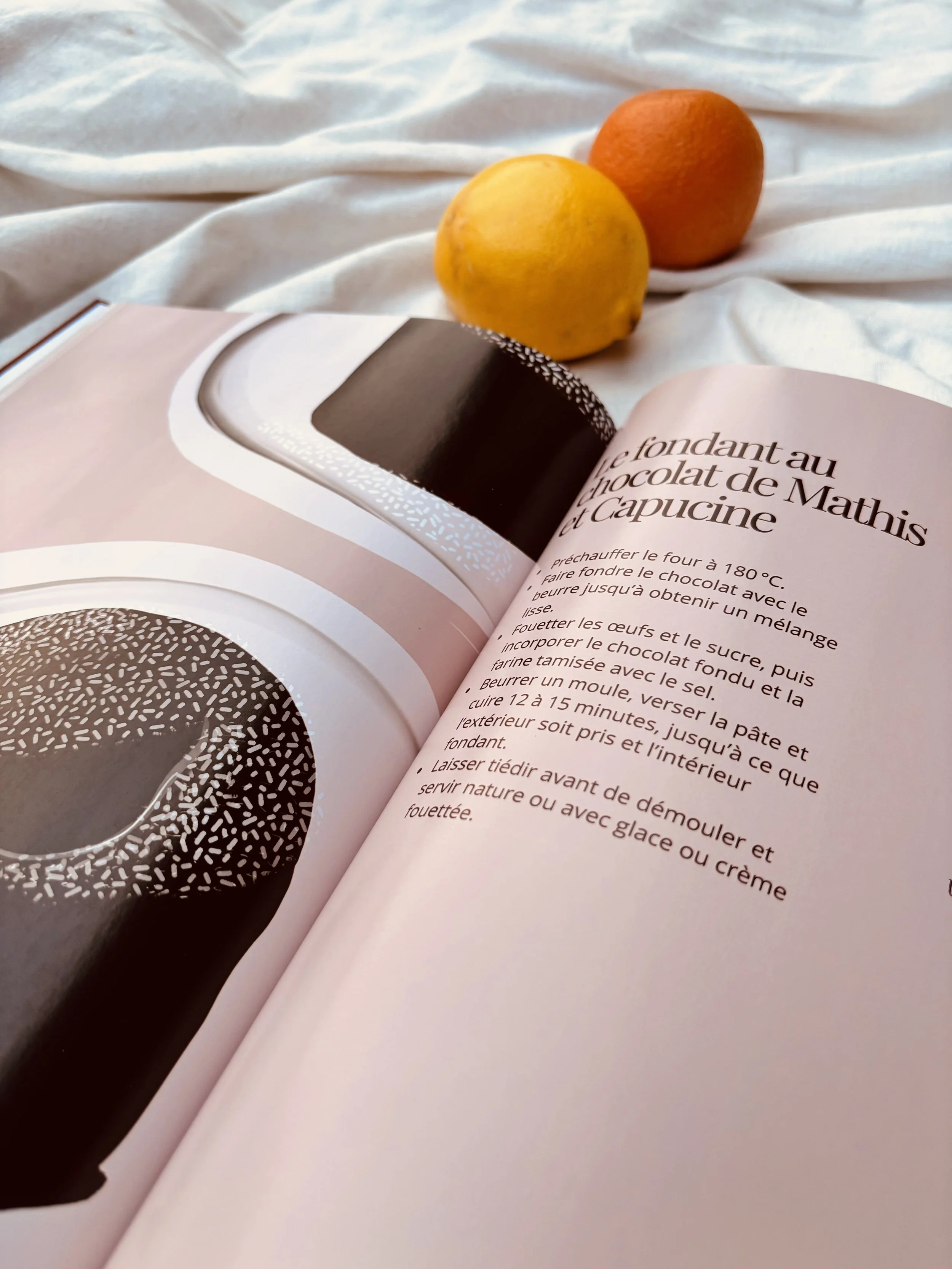 Un livre de cuisine ouvert montrant une recette de fondant au chocolat, deux agrumes (un citron et une orange) et un fond blanc en arrière-plan.