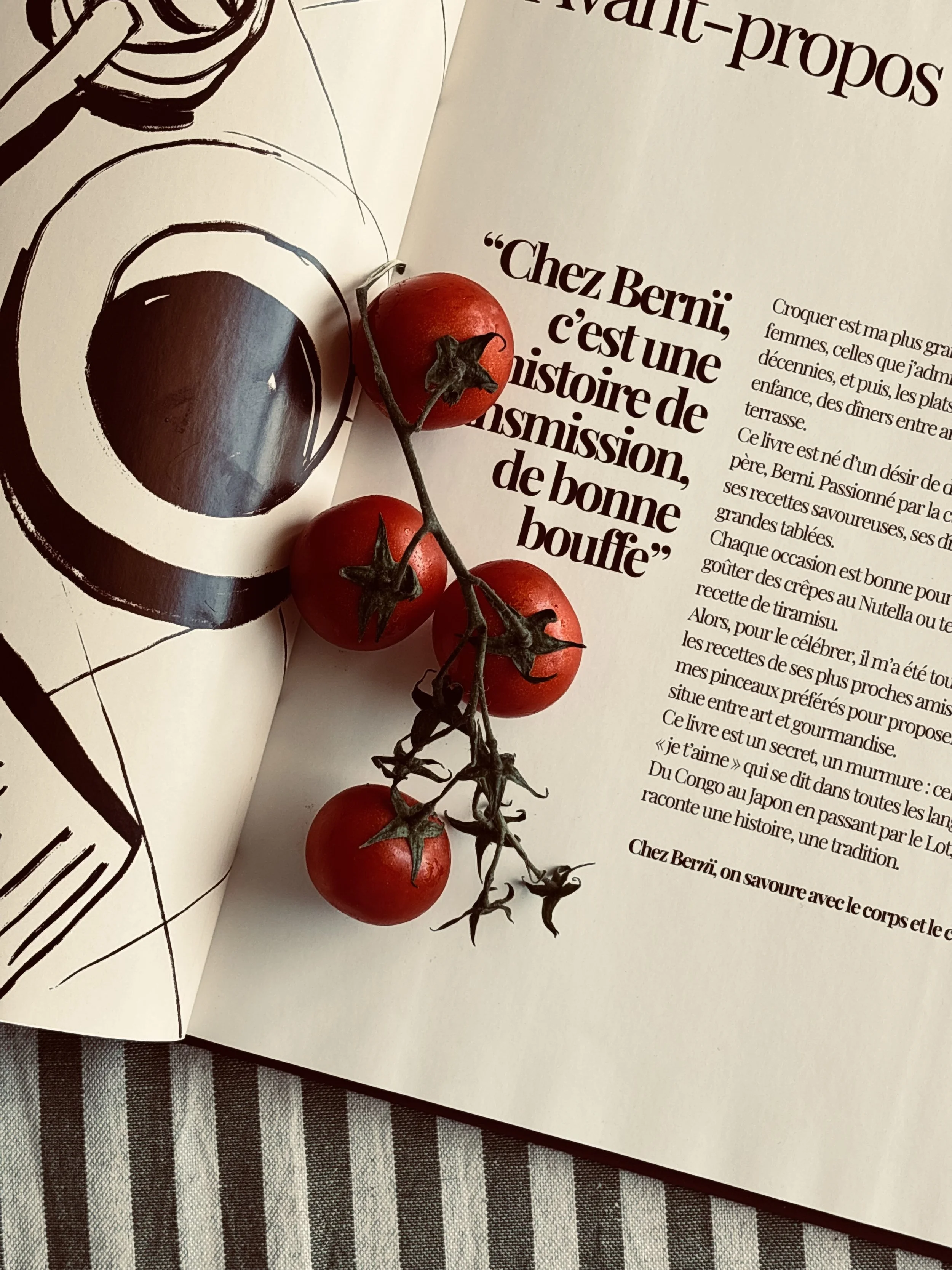 Une branche de tomates cerises rouges posée sur un livre ouvert avec du texte en français, à côté d'une illustration monochrome d'une tasse de café.