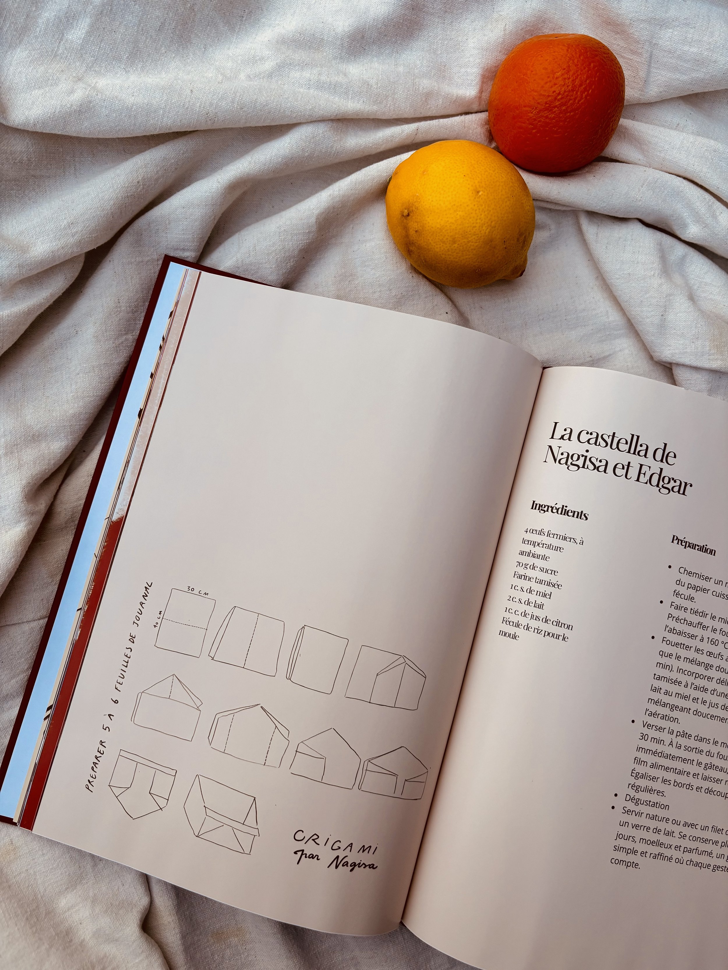 Une page d'un livre de cuisine avec des croquis de pliage d'emballages en papier, à côté de deux fruits, un citron et une orange, posés sur un drap blanc.