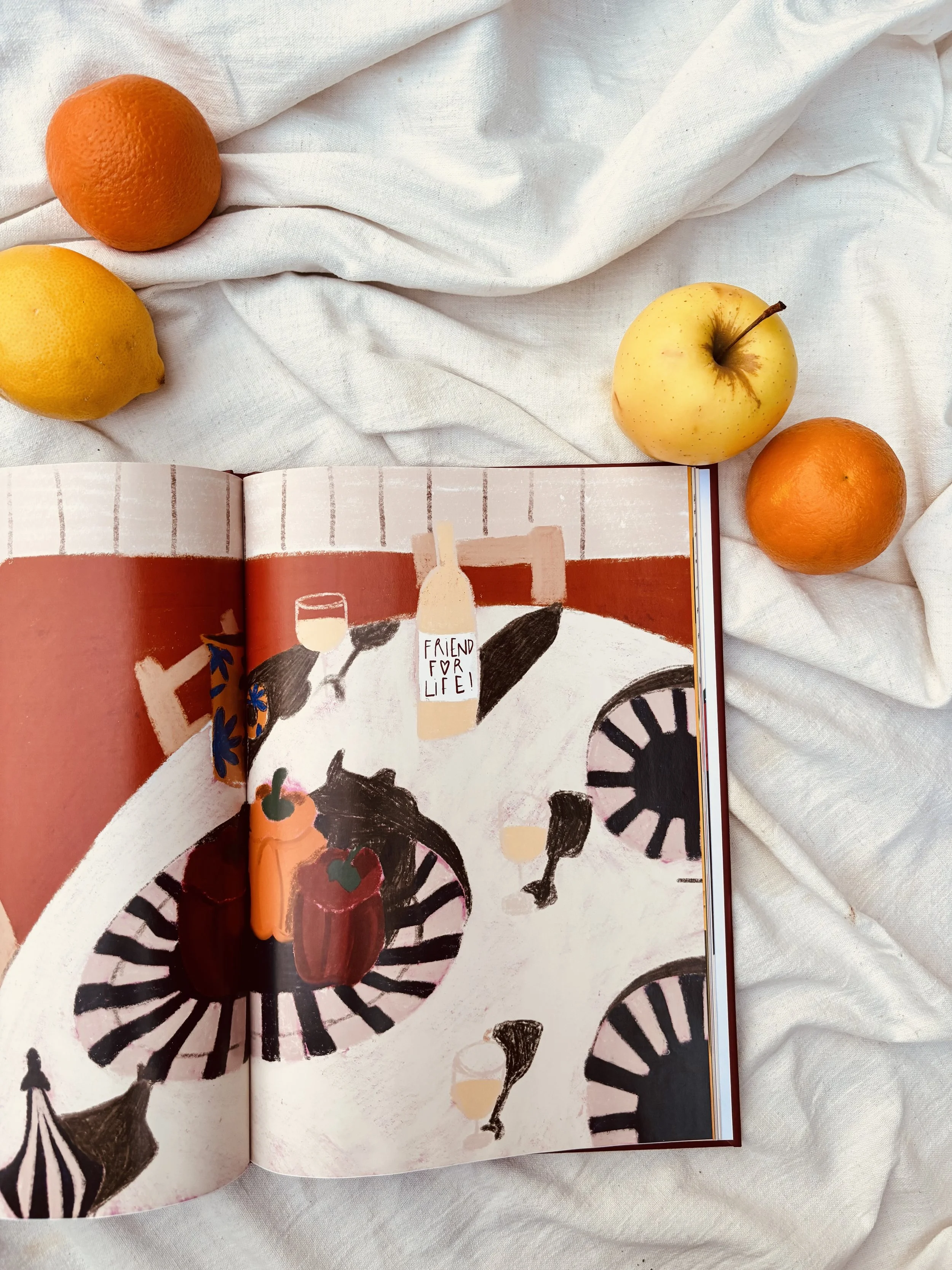 Un livre ouvert montrant une illustration abstraite d'une table avec des verres et une bouteille, entourée de fruits comme des oranges, un citron, et une pomme jaune, sur un tissu blanc.