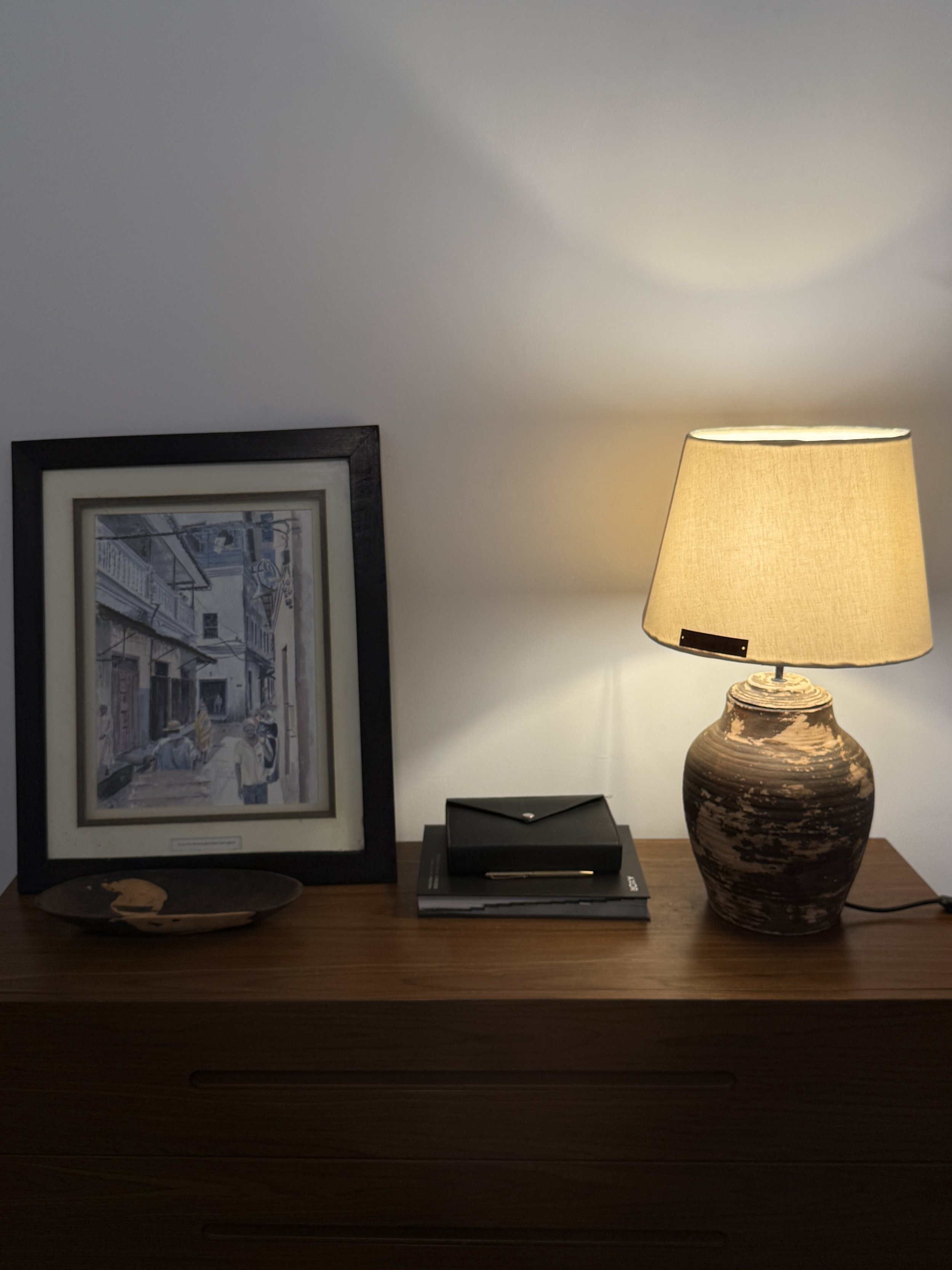 A wooden table with a framed black and white cityscape photograph, a decorative ceramic vase with a large beige lampshade, a black box, and a small decorative plate.