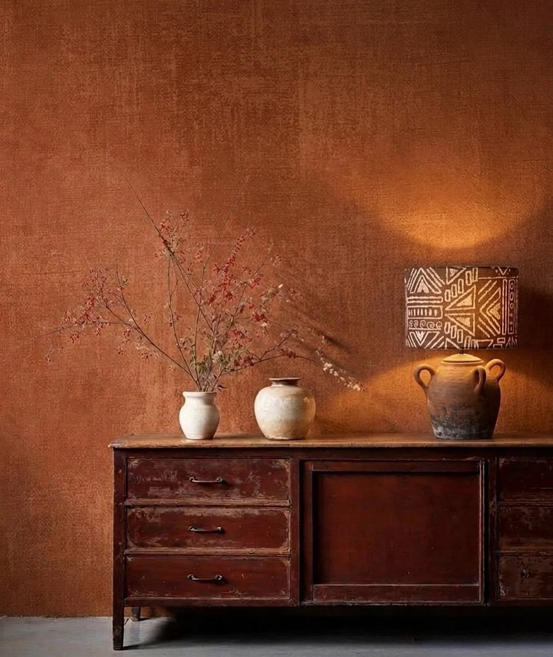 A wooden sideboard with three drawers and a large open space, topped with three vases, one holding branches with pink blossoms and another with a smaller round shape, beside a patterned table lamp, against a textured orange wall.