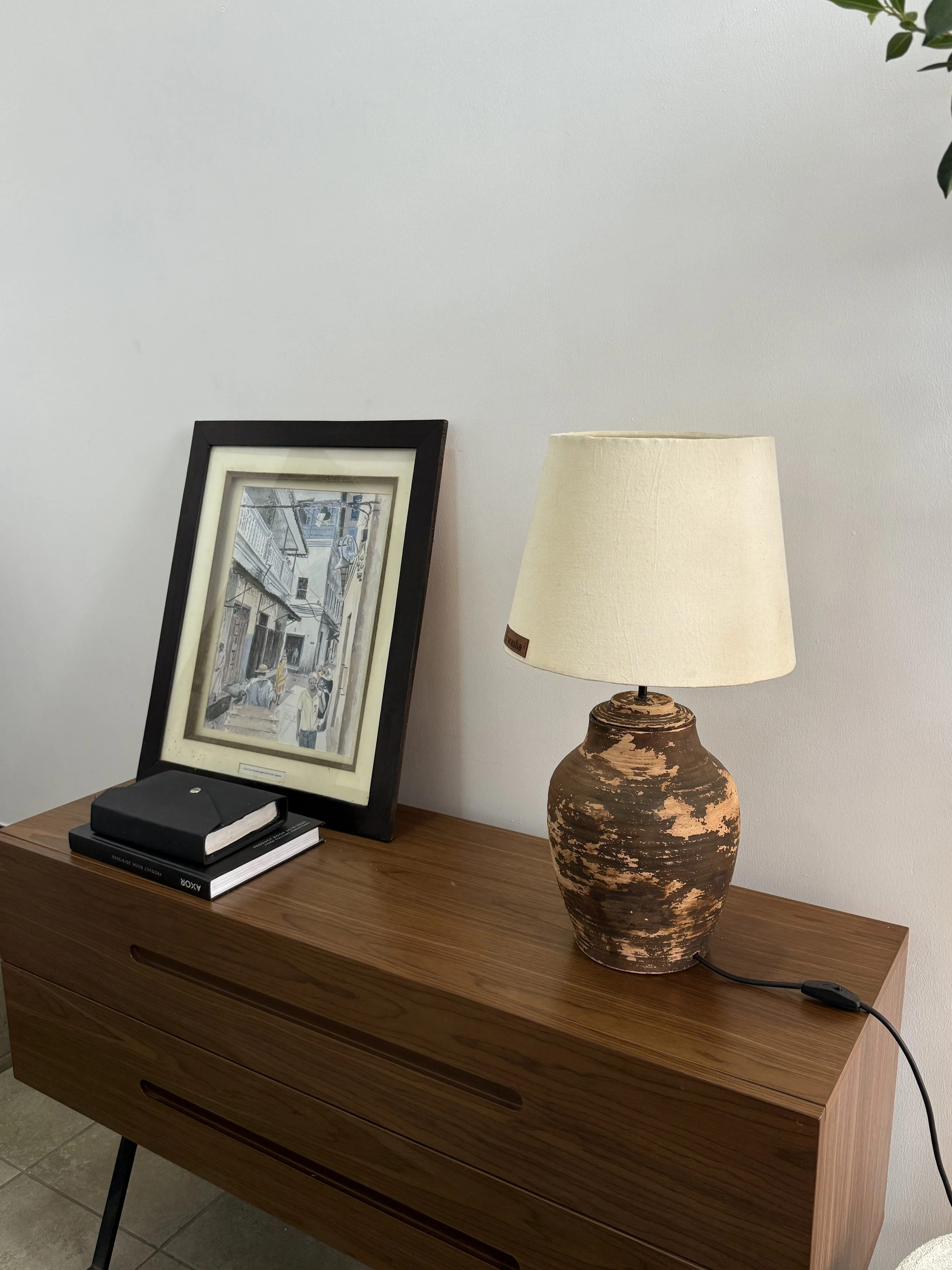 A wooden side table with a framed photo of a street scene, three books, and a black notebook on top. Next to the table is a large, rustic ceramic table lamp with a off-white lampshade. Part of a leafy plant is visible in the top right corner.