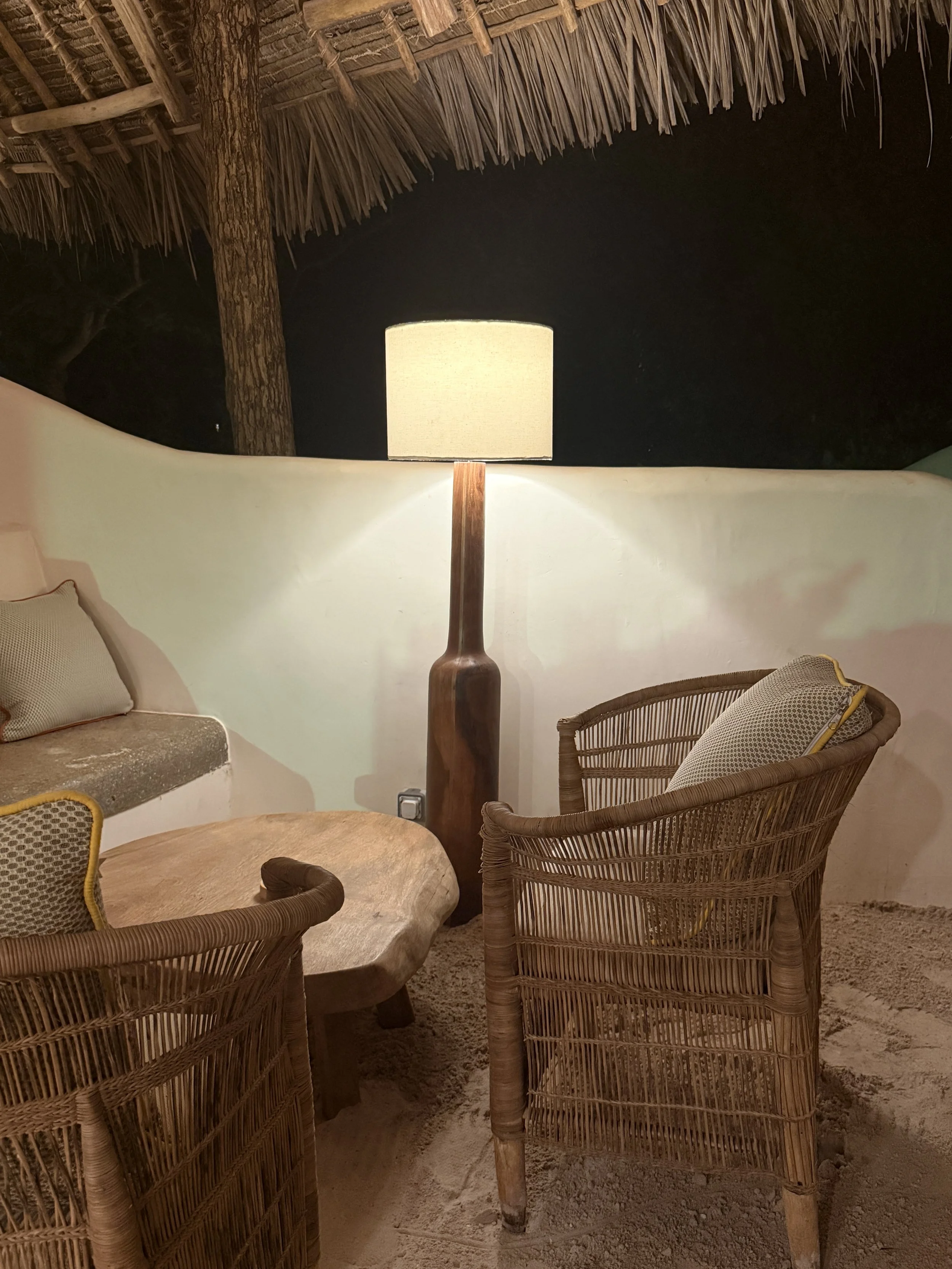 A cozy sitting area with wicker chairs, a wooden table, a lamp, and a thatched roof overhead, with sand or dirt on the ground.