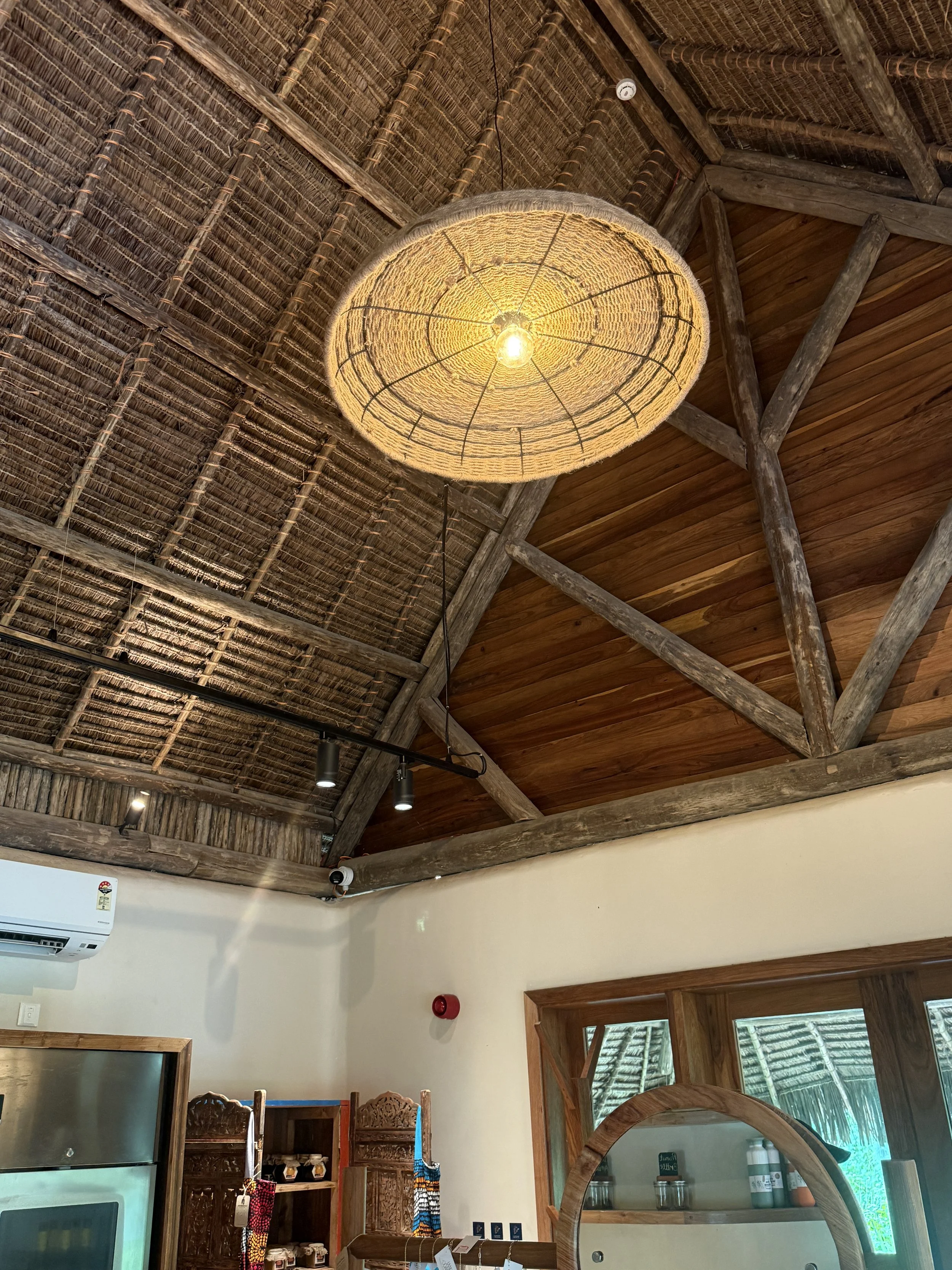 Interior of a rustic building with a thatched roof, wooden beams, and a hanging woven lamp with exposed light bulb. There are windows, a refrigerator, and decorative items on shelves.