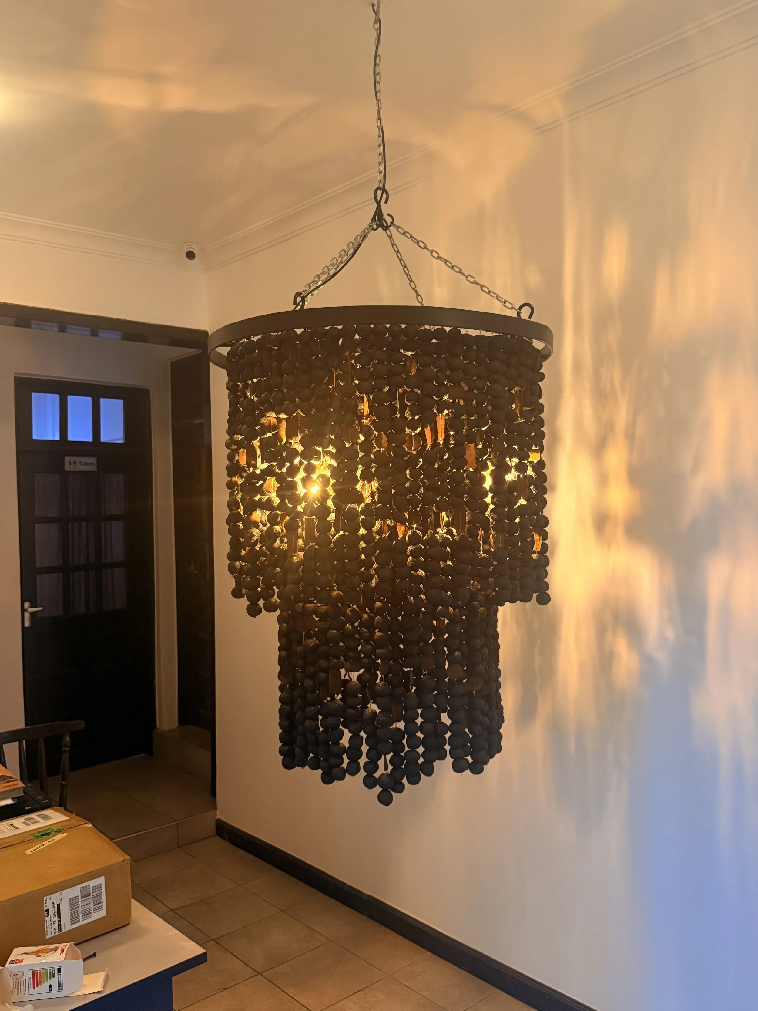 A decorative hanging chandelier made of small dark beads and wooden pieces, illuminating a room with warm light.