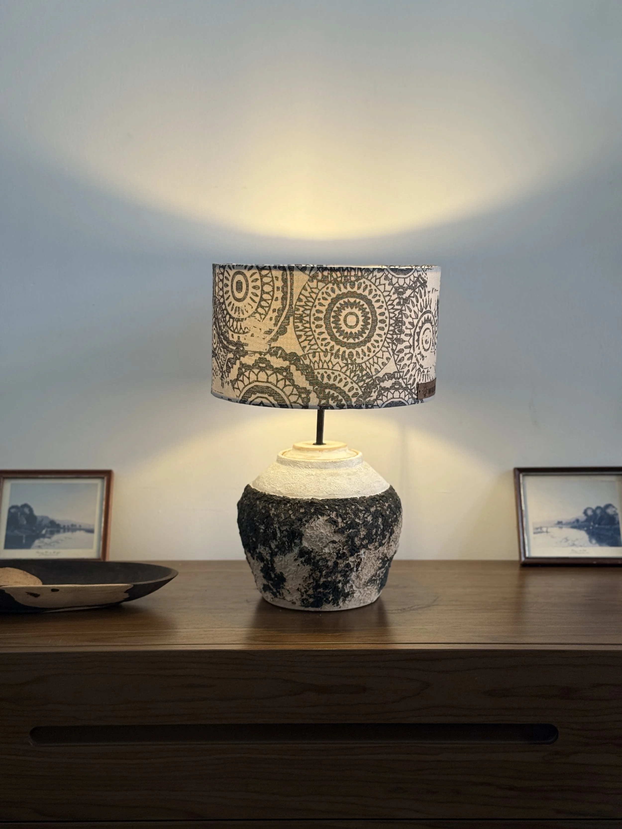 A table lamp with a cylindrical shade featuring black and white geometric and circular patterns, positioned on a wooden surface with framed photos and a decorative dish in the background.