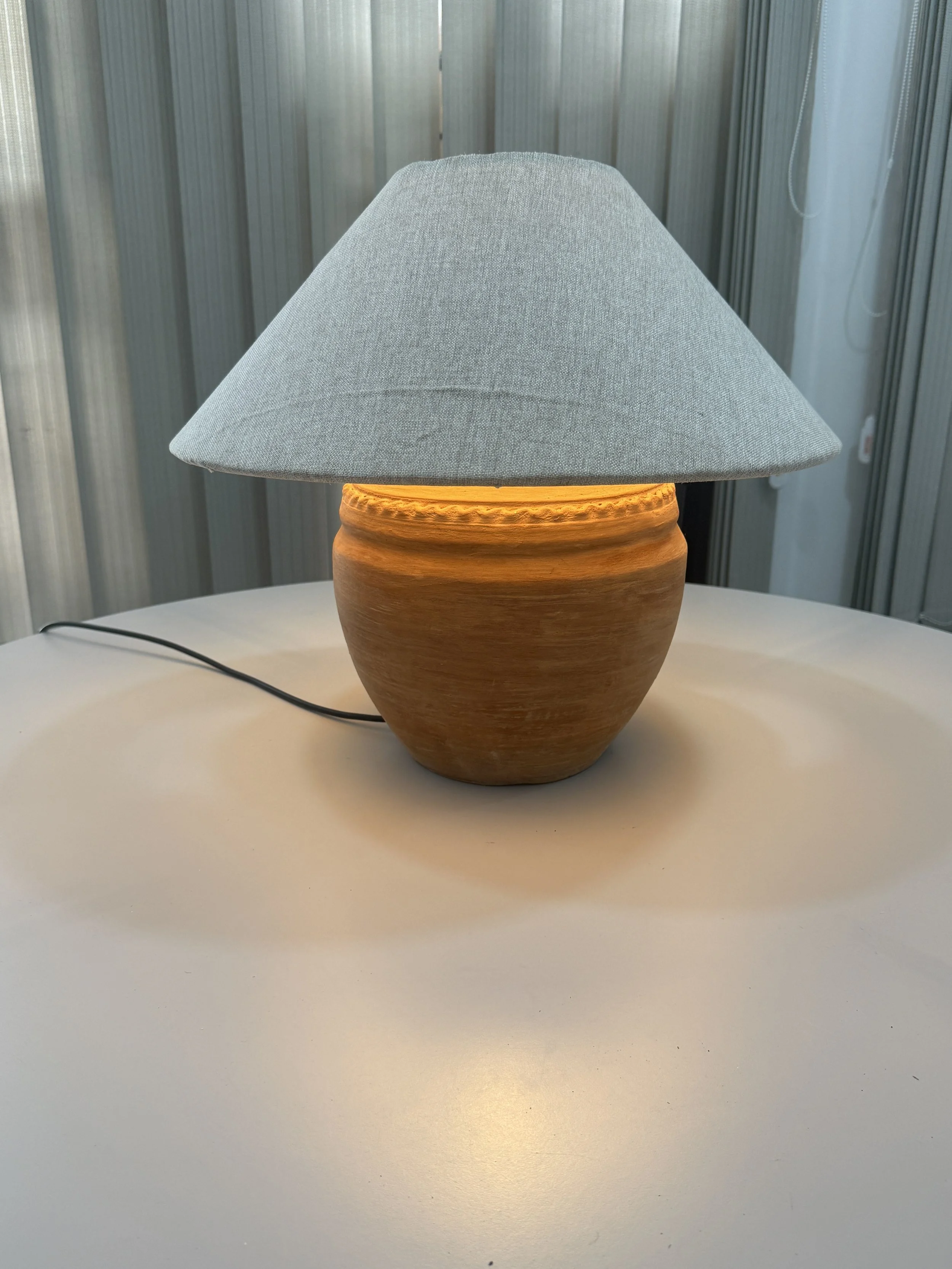 A table lamp with a gray fabric shade and a ceramic base, placed on a white surface with a black cord, in front of a metallic-paneled background.
