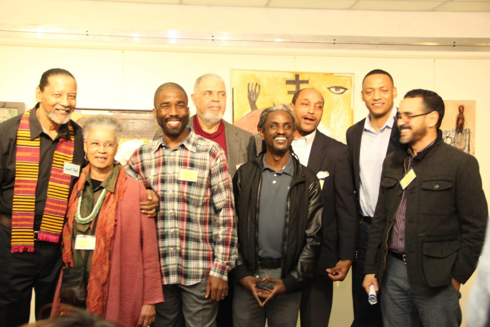 Group of nine diverse people smiling and posing for a photo at an indoor event, with art on the wall behind them.