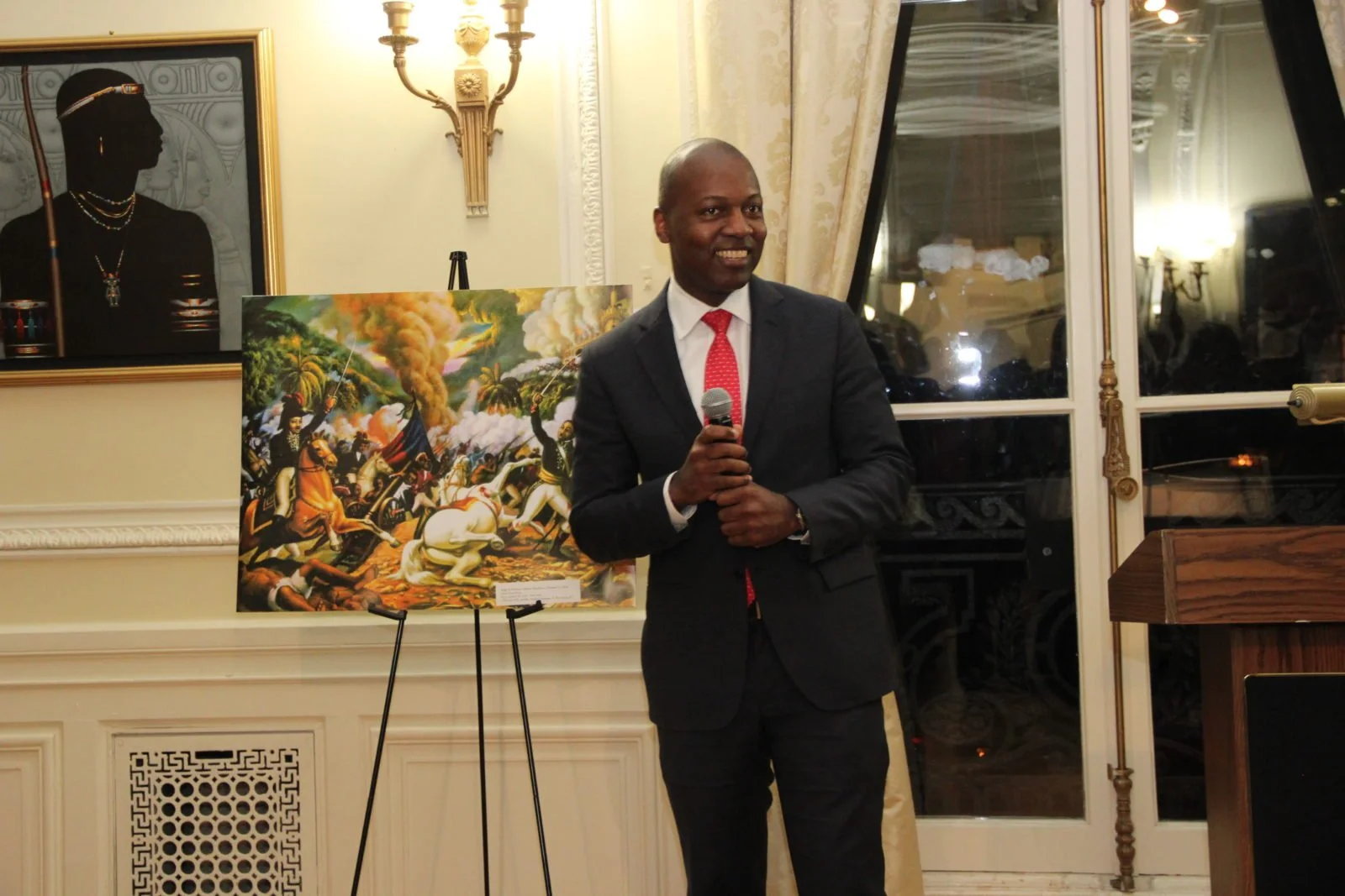 A man in a black suit holding a microphone, standing next to a colorful painting of a battle scene with horses and soldiers in a well-lit room.