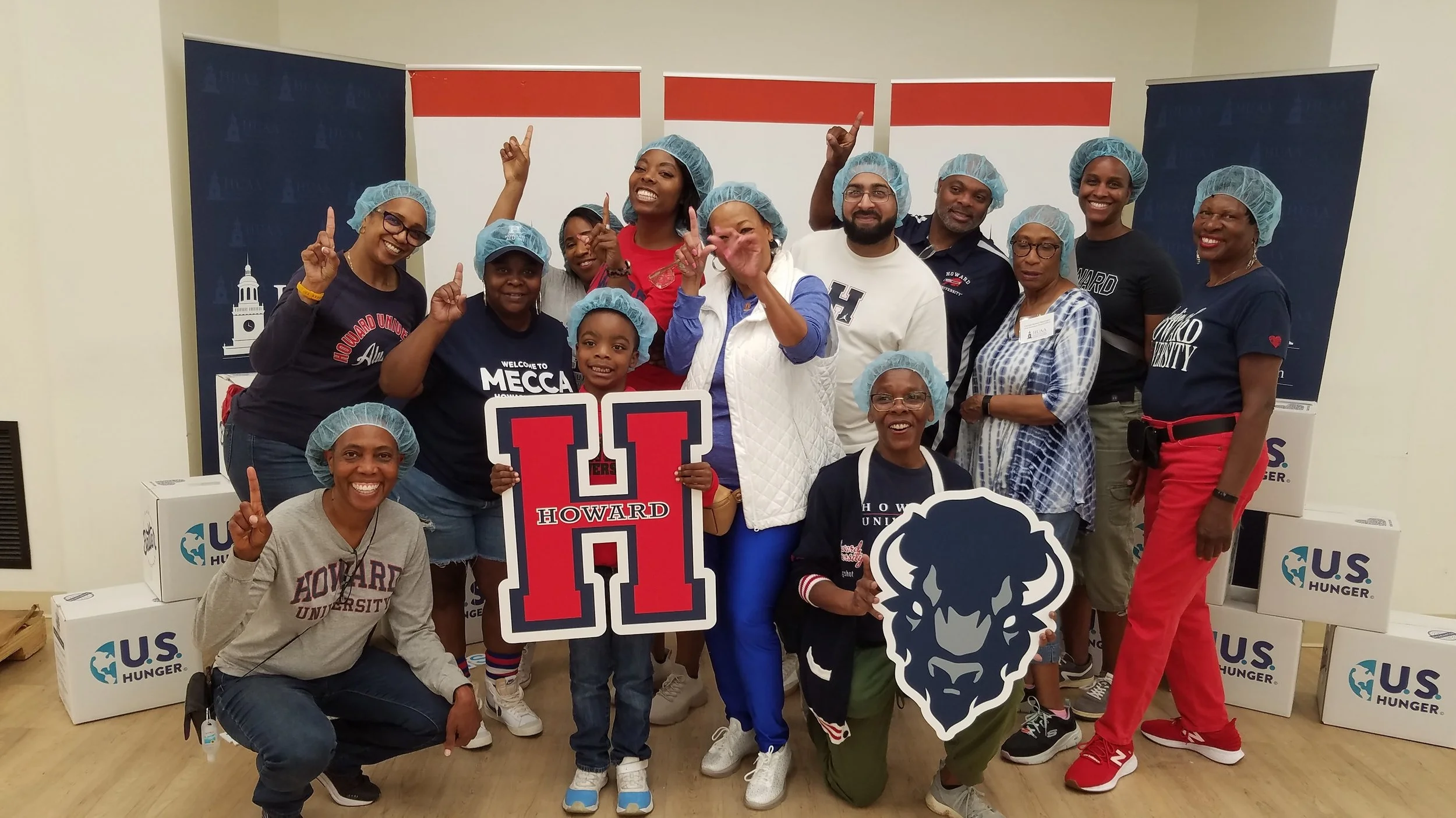Group of people wearing hairnets, posing with signs and smiling, indoors at a community event with U.S. Hunger boxes in the background.