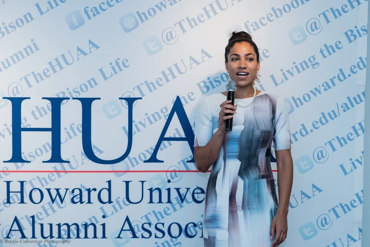 A woman holding a microphone speaking at an event with a backdrop displaying 'Howard University Alumni Association,' social media handles, and the hashtag '@TheHUAA'.