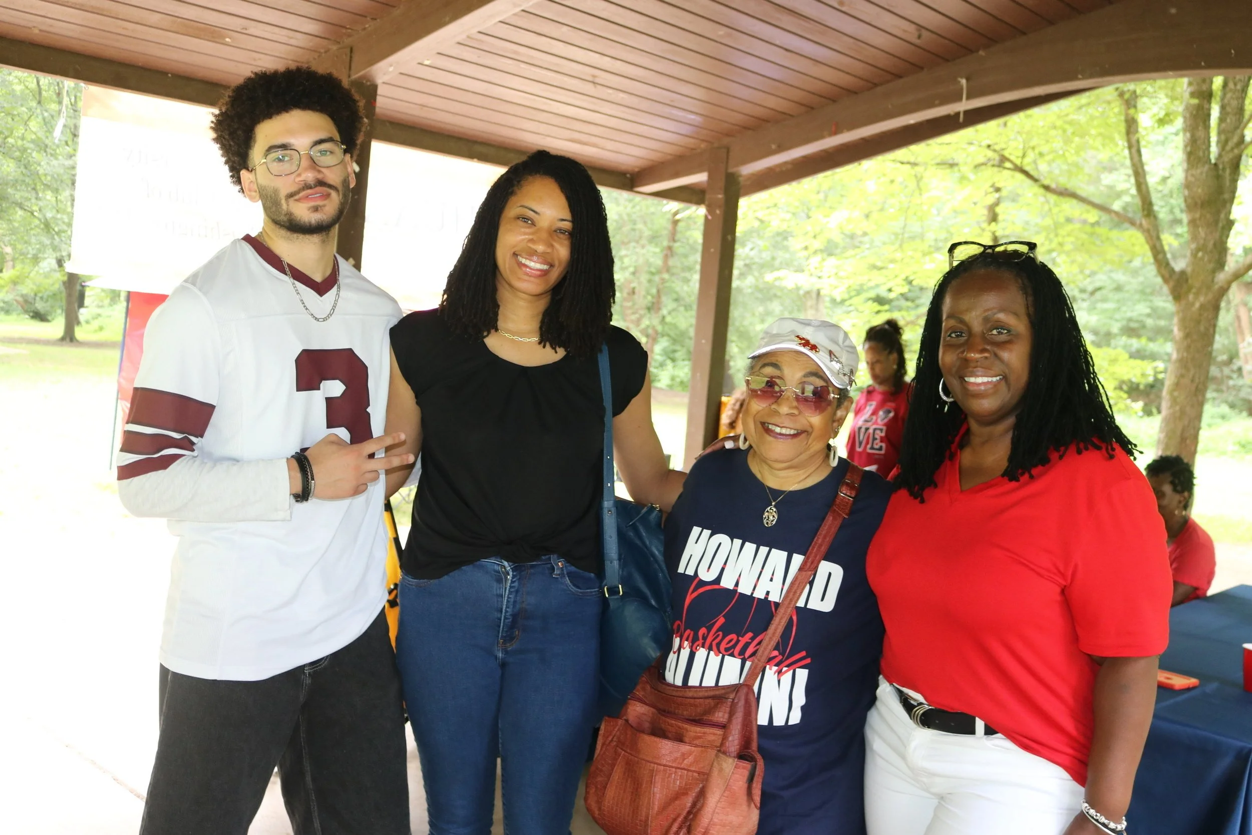 Four women and one man smiling and standing together outdoors under a pavilion, with trees and other people in the background.