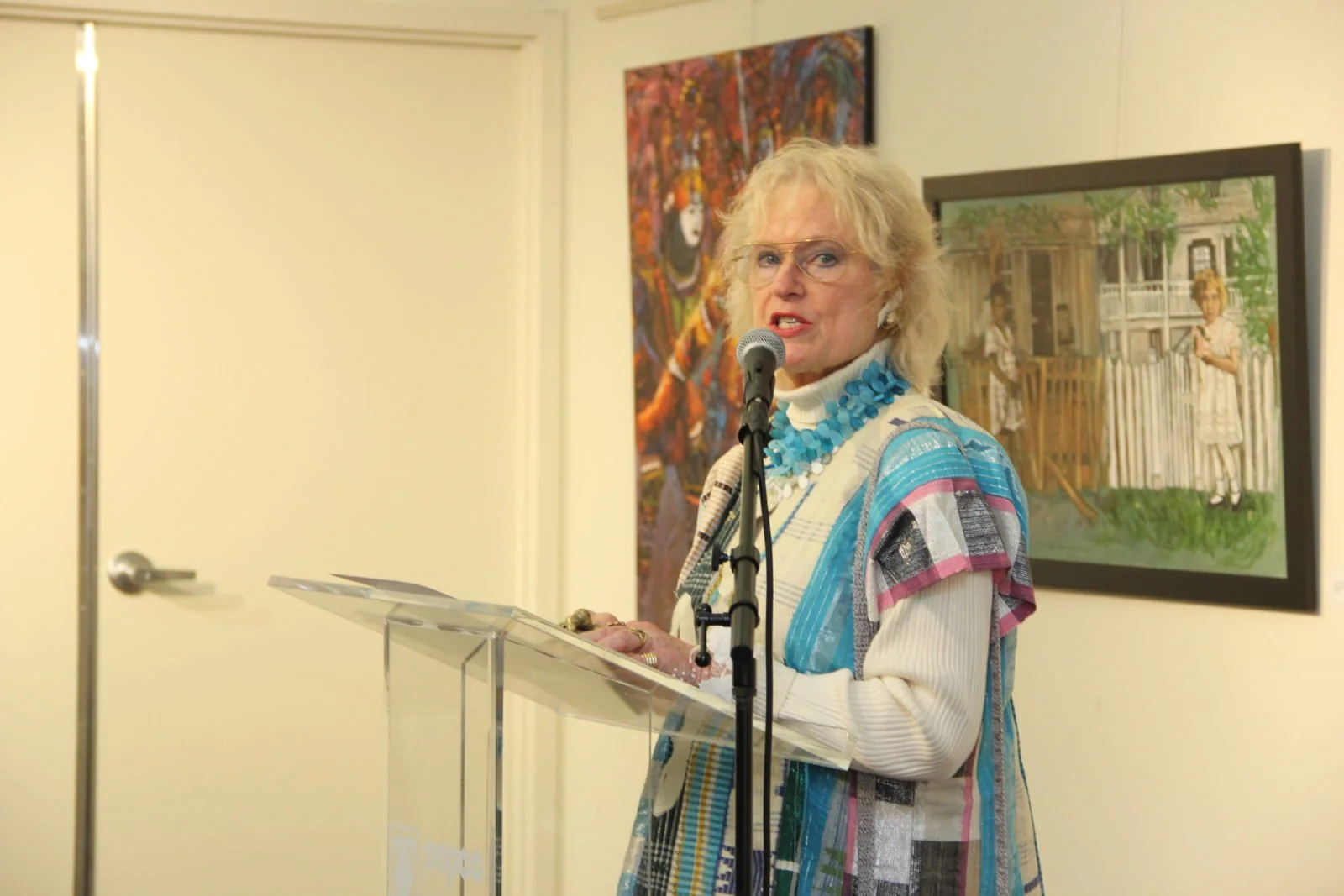 An older woman with blonde curly hair and glasses speaking into a microphone at an art gallery event, wearing a colorful, patterned cardigan with a blue necklace. Behind her are two framed paintings, one depicting children on a porch and the other a 