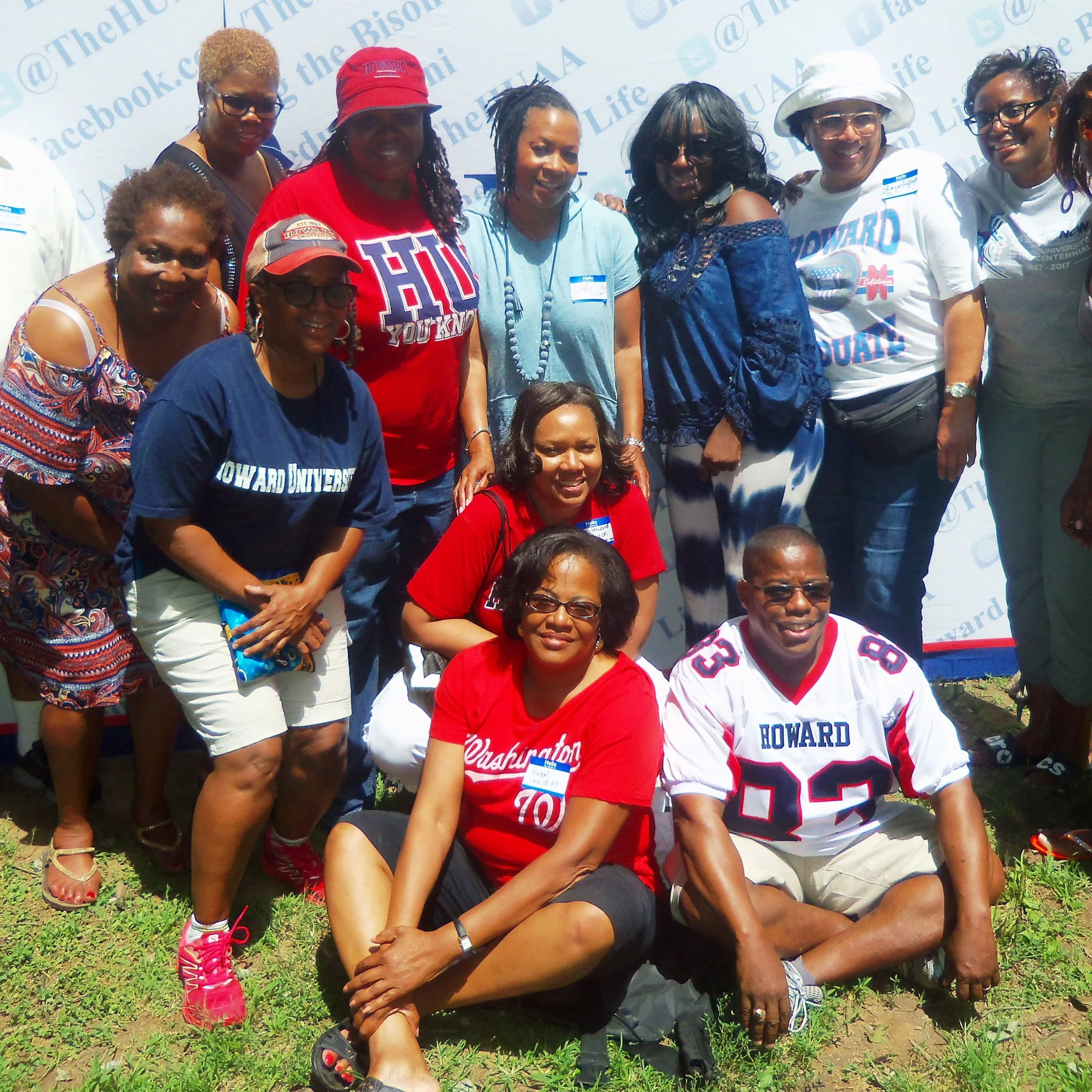 Group of adults posing outdoors with a Howard University backdrop, some wearing Howard gear, during a daytime event.