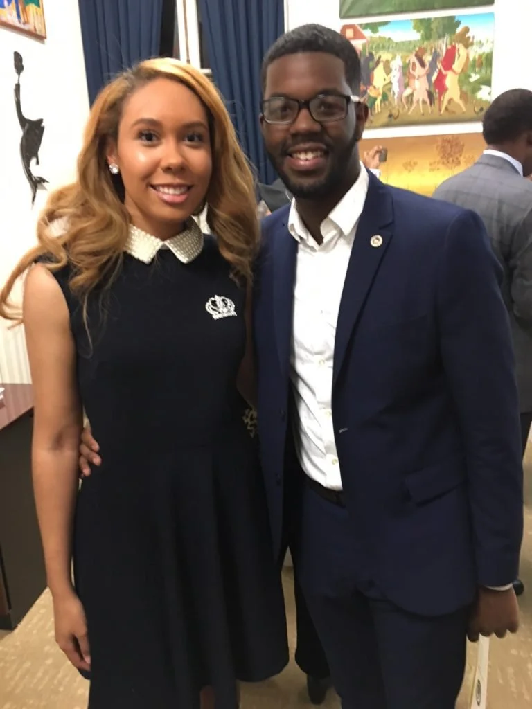 A woman in a black dress with a white pearl collar and a man in a dark blue suit and glasses, both smiling, standing indoors at an event with paintings on the wall behind them.