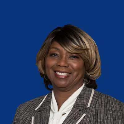 Portrait of a smiling woman with short, blonde-highlighted hair, wearing a gray checkered blazer and white blouse, against a blue background.