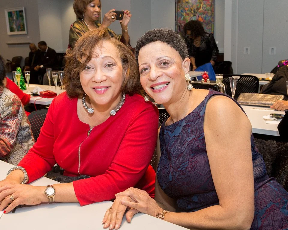 Two women are sitting at a table, smiling and posing for a photo at a social event or gathering. They are dressed nicely, with one in a red top and the other in a dark blue dress. The background shows other people, some seated and some standing, some