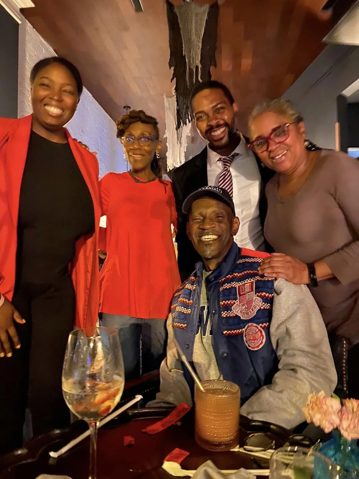 A group of six people smiling and posing at a social gathering or restaurant. Five are standing, one is seated. They appear joyful, with a table in front holding a glass, a drink, and some flowers.