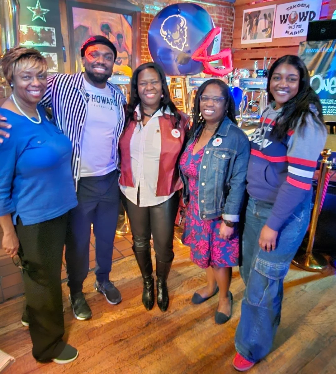 Group of six people standing inside a bar or restaurant, smiling for a photo, with balloons and neon signs in the background.