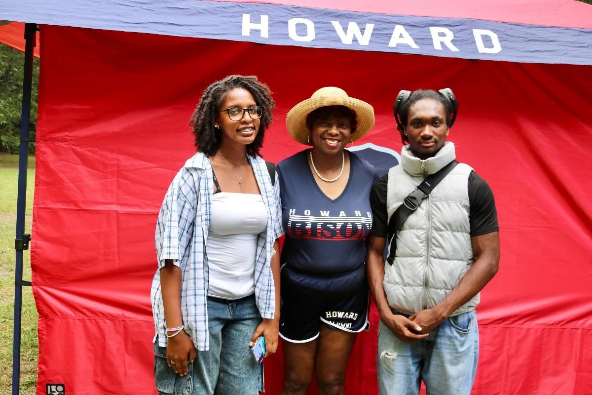 Three people standing together in front of a red Howard University banner outdoors. The person in the center is a woman wearing a straw hat, a blue Howard shirt, and black shorts. The person on the left is a woman with glasses, wearing a white top an