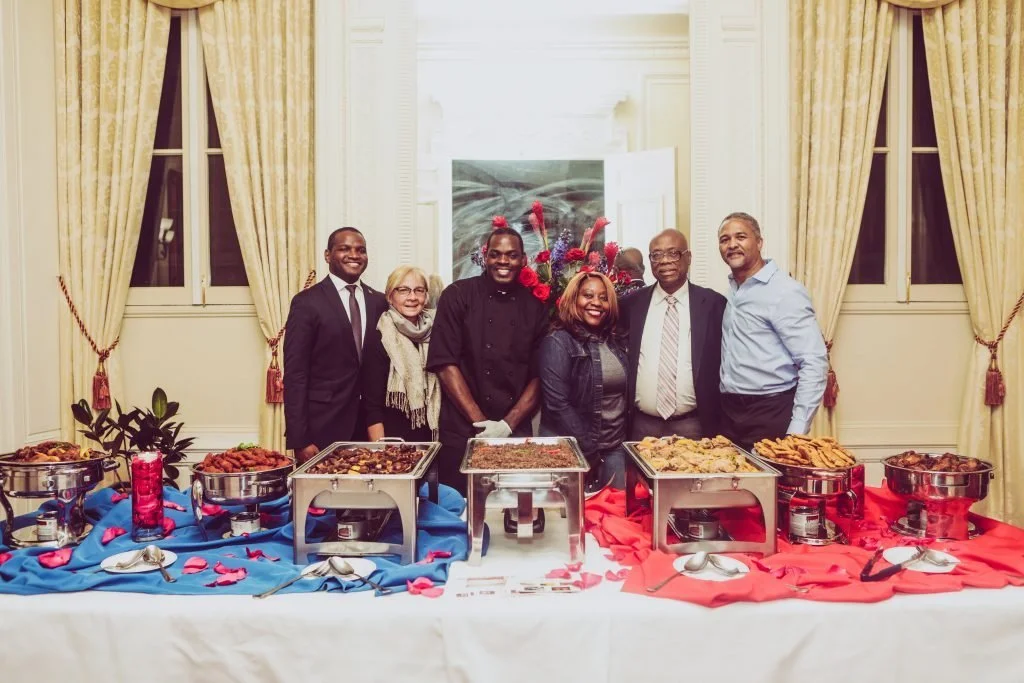 Group of six people standing behind a buffet table with various dishes at a banquet or celebration in an elegant room with tall windows and cream-colored curtains.