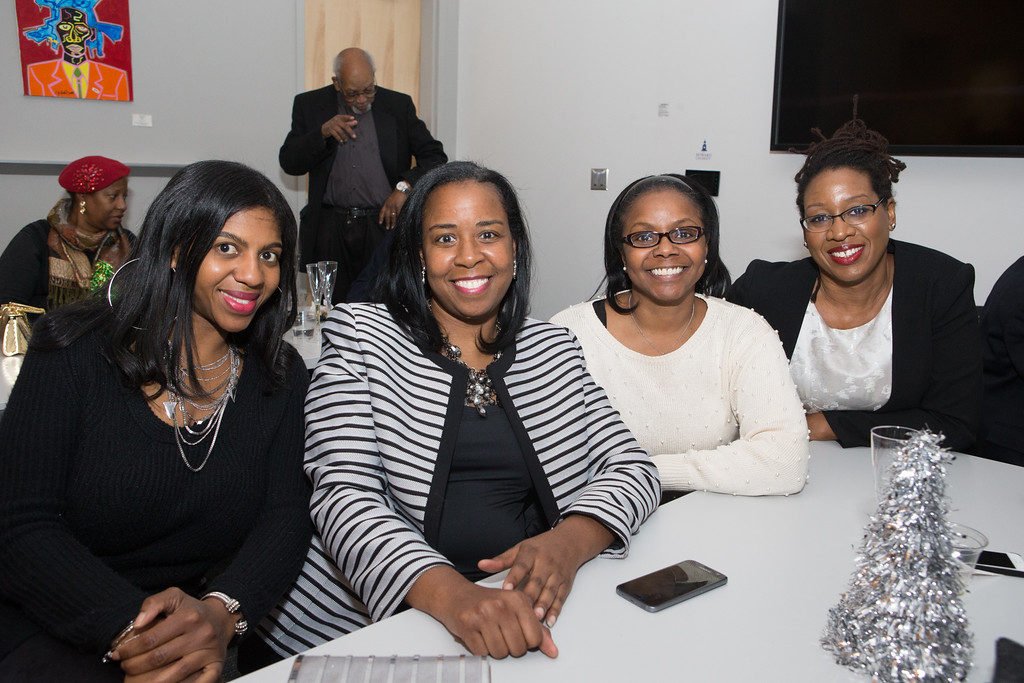 Four women sitting at a round table smiling for the camera at a social gathering or event, with a small silver Christmas tree decoration on the table.