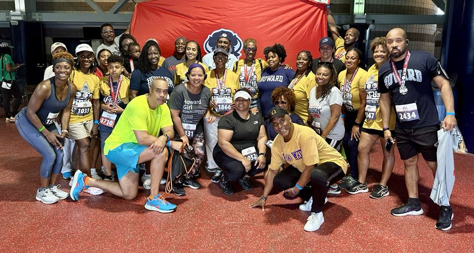 Group photo of diverse men, women, and children at a running event wearing athletic clothes and race bibs, indoors with a red carpeted floor and a large red and blue banner or flag in the background.