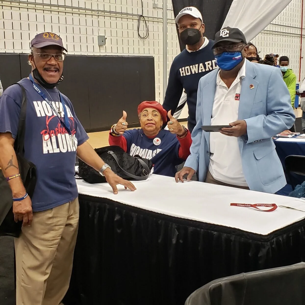 A group of four people, three men and one woman, standing around a table at an indoor event. They are all wearing clothing supporting Howard University, and three are wearing masks. The woman in the center is giving a thumbs-up and smiling.