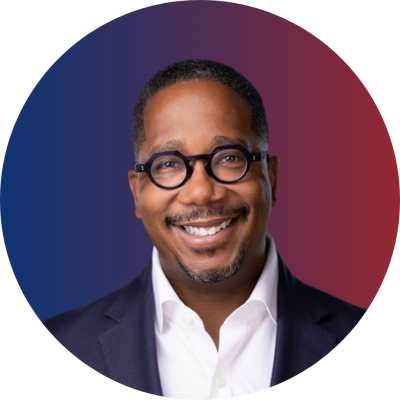 Smiling man with glasses in a suit and white shirt, standing in front of a background with blue and red gradient colors.