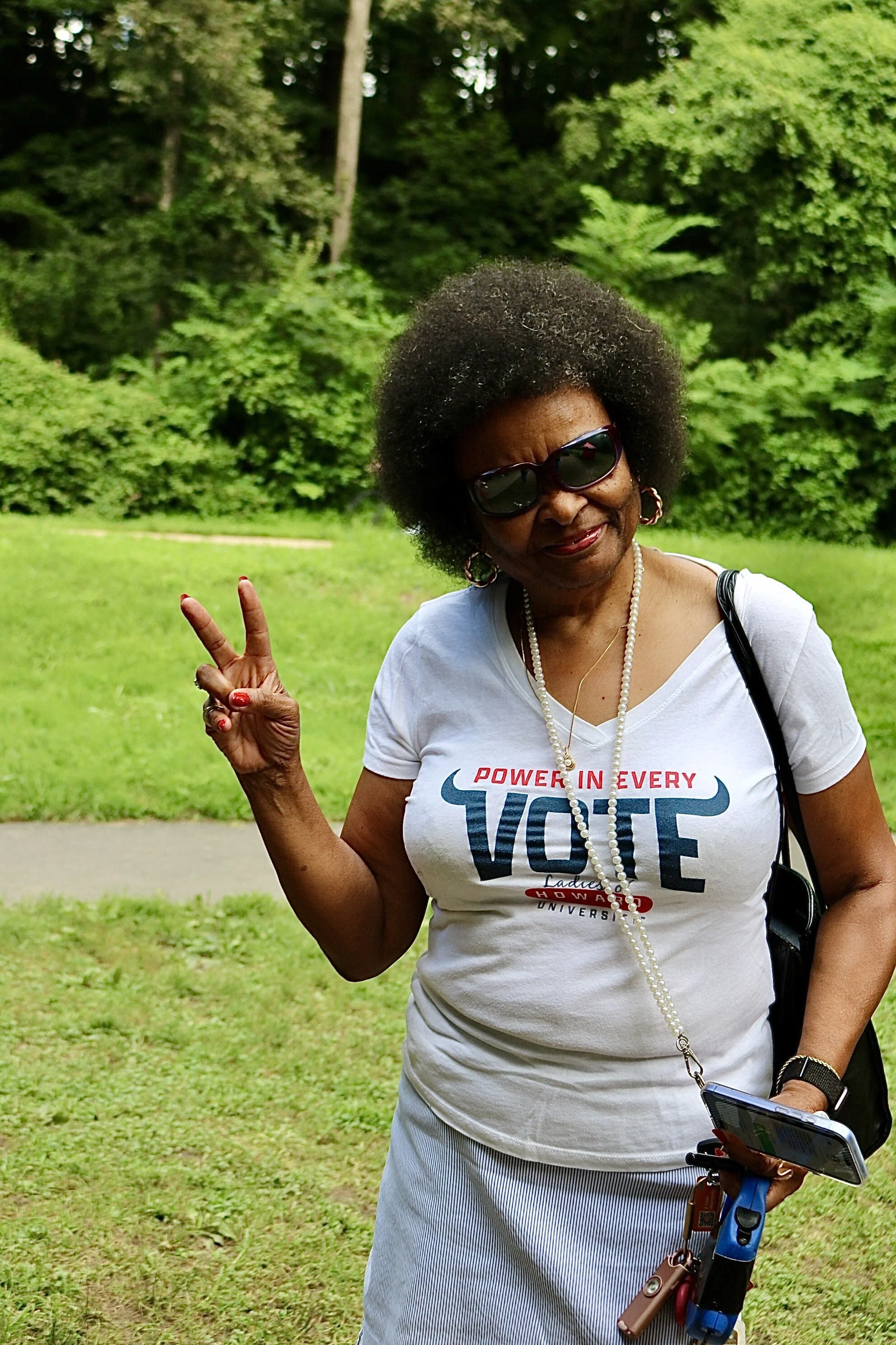 A woman wearing sunglasses and a white T-shirt with the slogan 'Power in Every Vote' is standing outdoors on a grassy area with trees in the background. She is making a peace sign with her right hand, smiling, and holding a phone and keys in her left