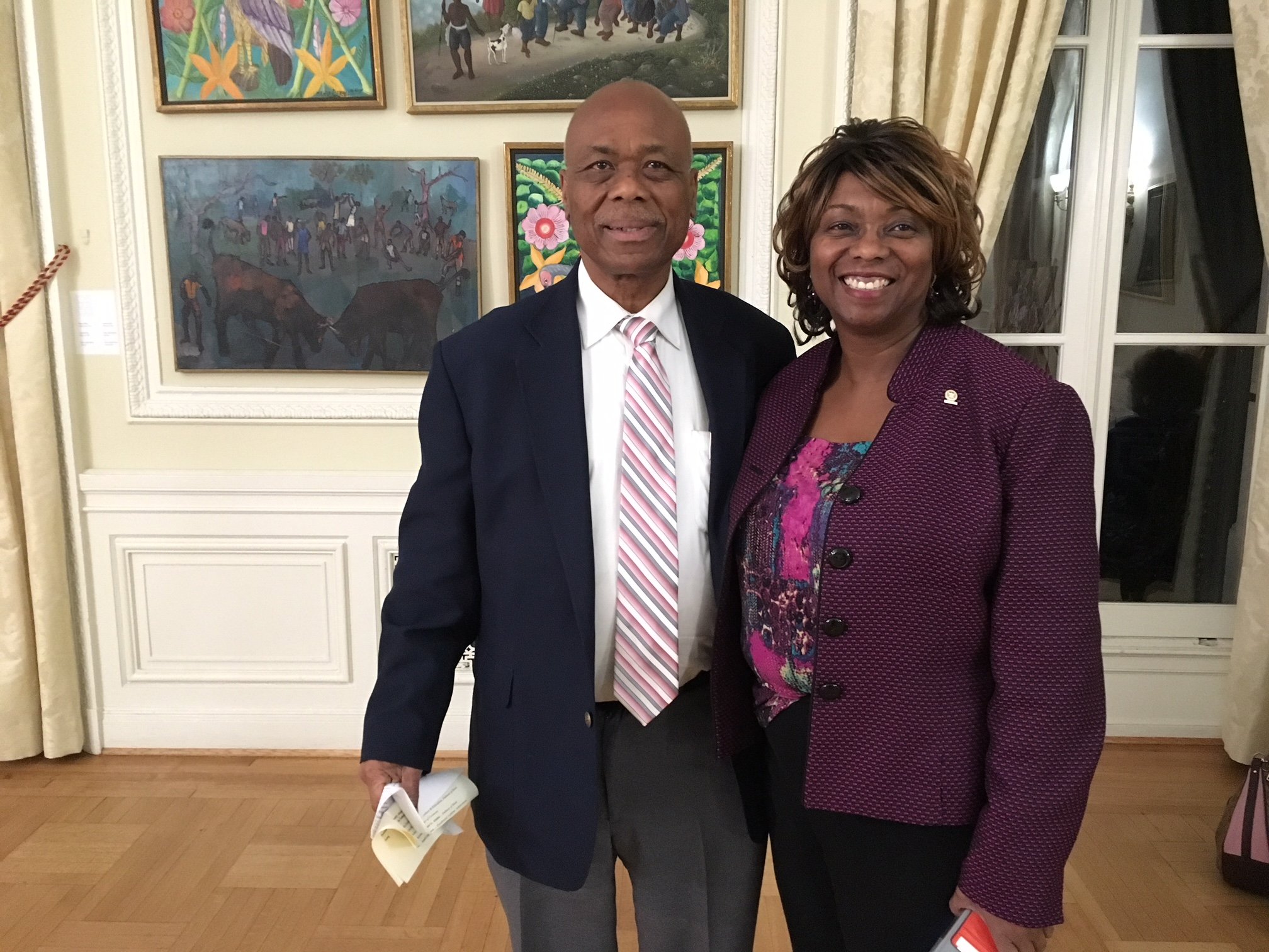 A man and woman smiling and posing together in a room with artwork on the wall and a window behind them. The man is wearing a dark suit with a striped tie and the woman is wearing a maroon blazer with a colorful blouse underneath.
