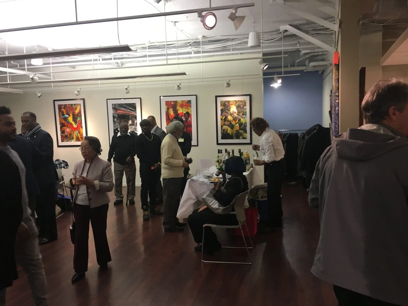 A group of diverse people mingling at an art gallery event with colorful artwork hanging on the walls and a table with drinks and snacks.