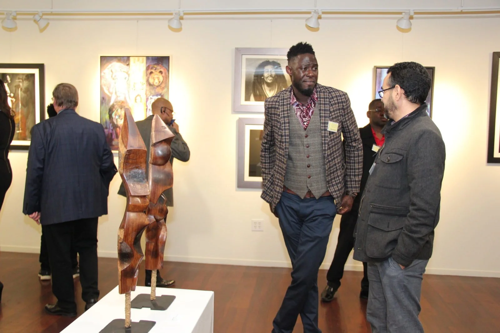 Two men are talking in an art gallery, surrounded by paintings and sculptures, with other visitors in the background.
