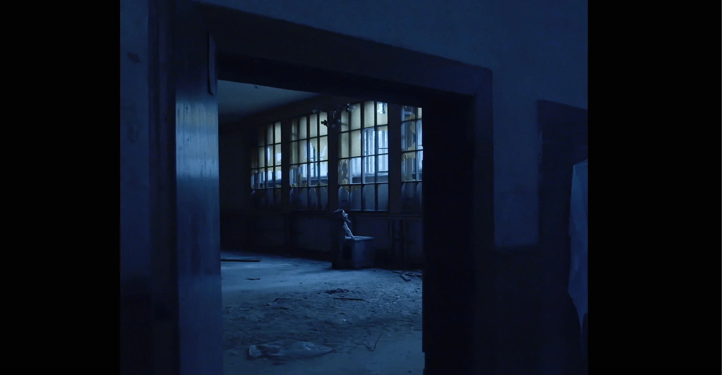 Dark interior of an abandoned building with a large window, broken glass, and a dilapidated floor. A figure is sitting on a bench near the window, looking out. The scene is dimly lit with blue tones, suggesting nighttime.