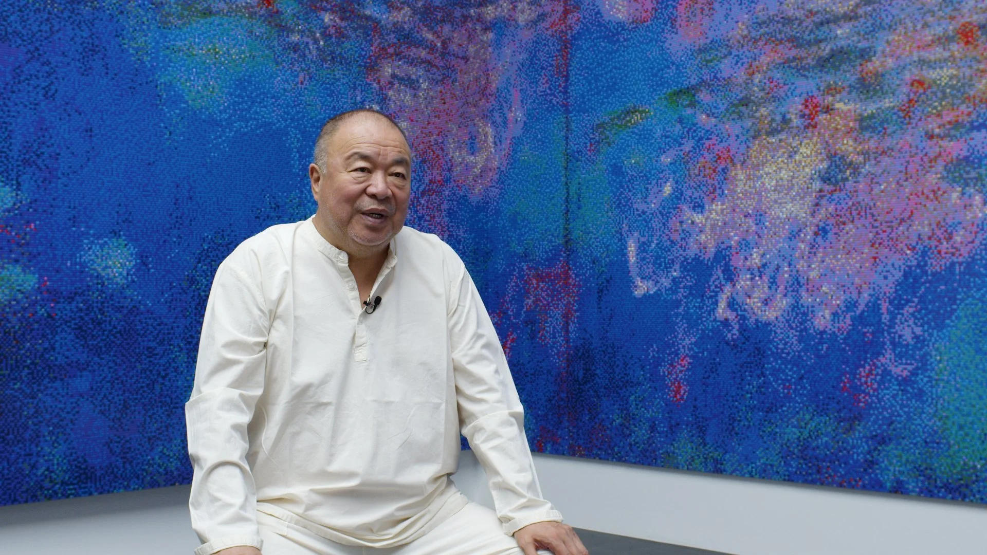 An older man wearing a white shirt sitting in front of a large colorful pixelated artwork on a wall.