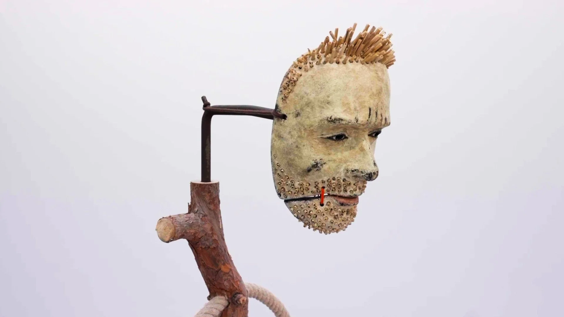 Art installation featuring a human face sculpture with a branch neck, wore with a head of wooden sticks, displayed against a plain white background.