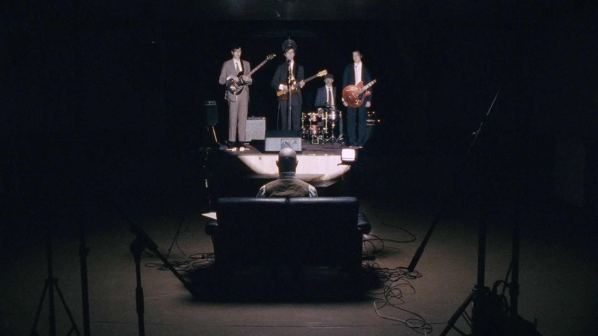 A band performing on a small stage in a dark room, with a drummer, and three guitarists, and an audience member watching from the front.