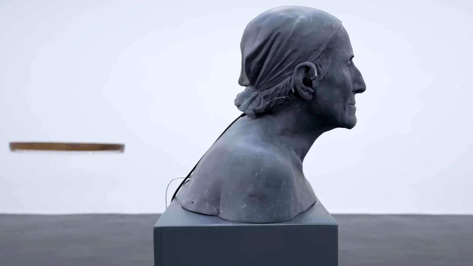 Bronze bust sculpture of a woman with hair tied back, displayed on a pedestal against a cloudy sky.