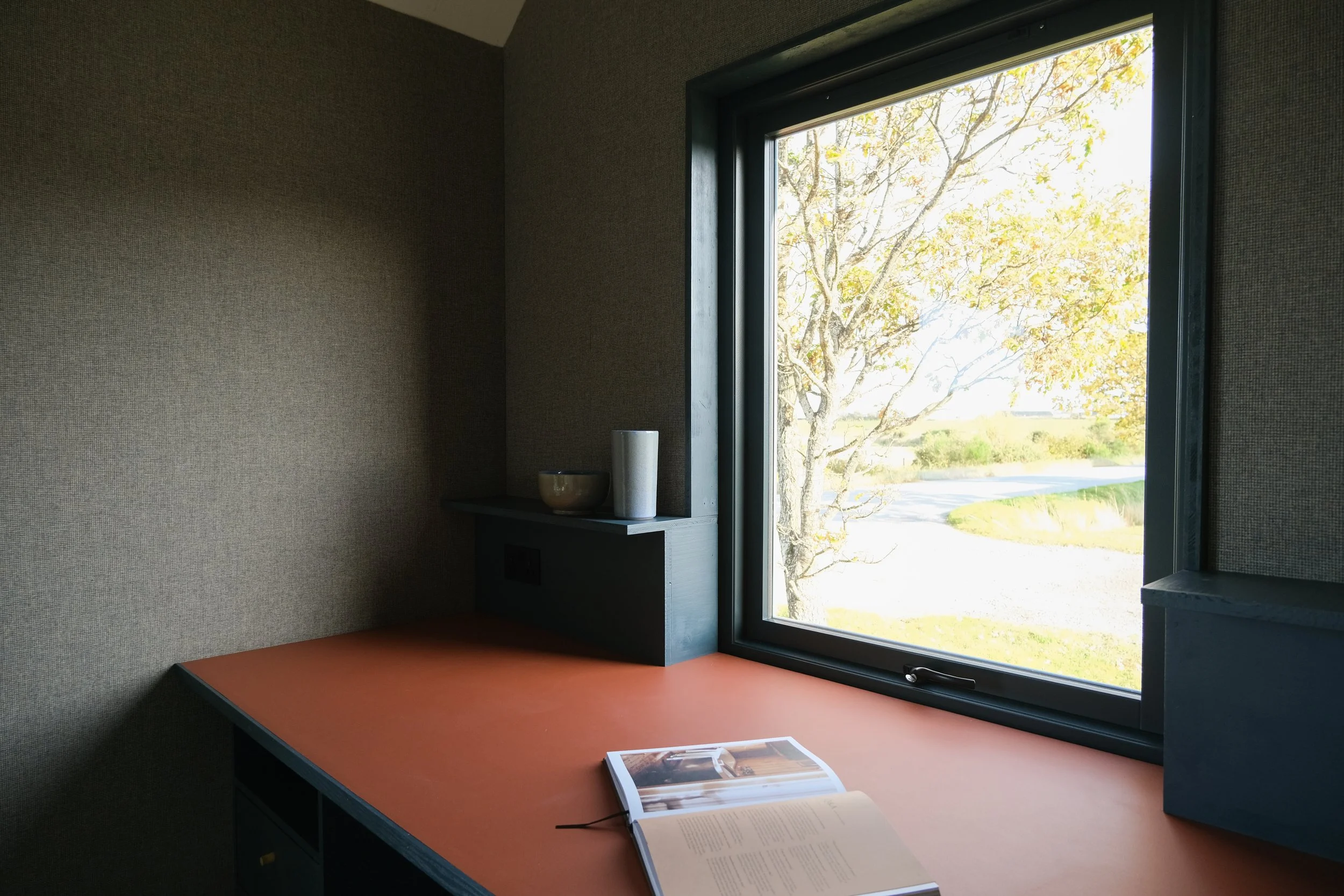 Interior of a room with a desk near a large window showing an outdoor landscape with trees and a road.